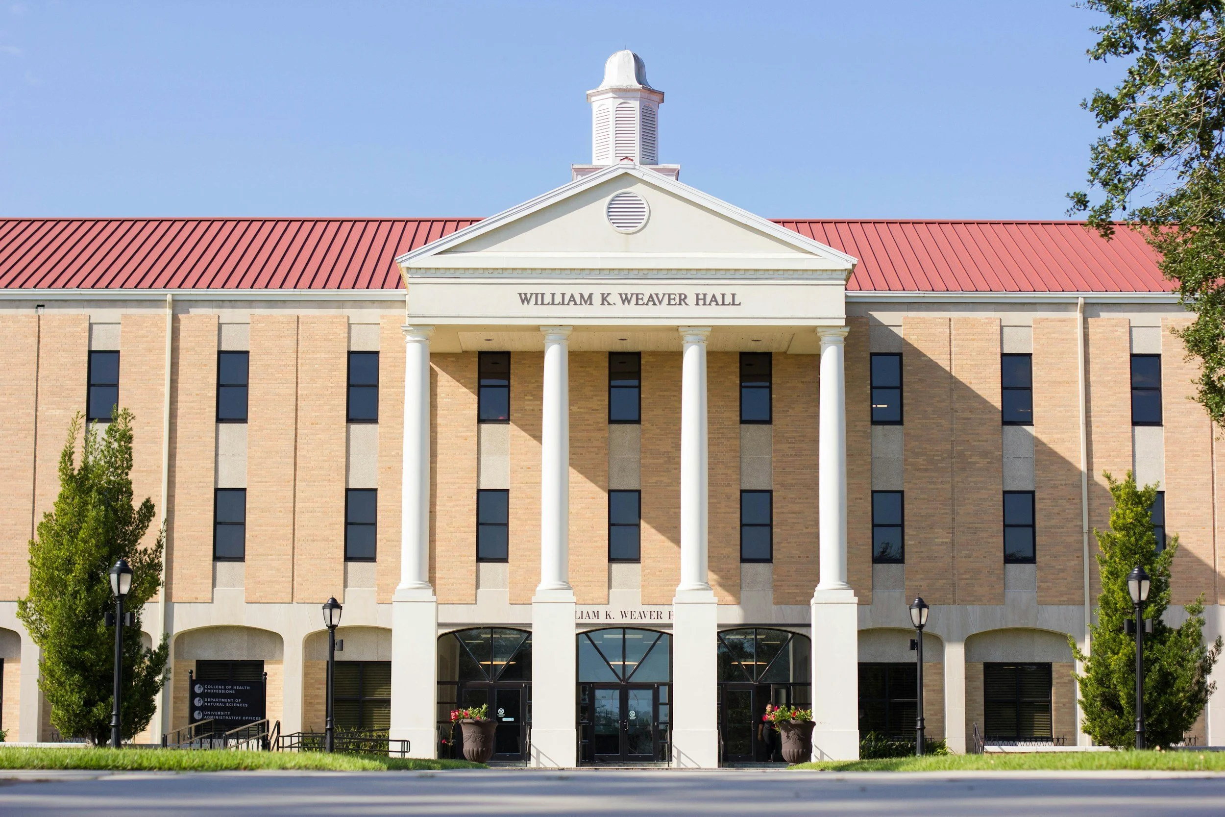 Front view of William K. Weaver Hall, a courthouse with four white columns, brick facade, red roof, and a small clock tower, with trees and lamp posts in front.