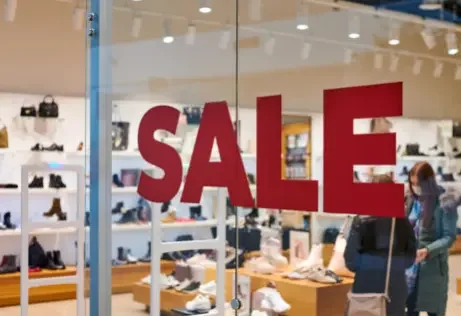 Store window with a large red 'SALE' sign, showcasing shoes and handbags on shelves inside the store, with customers browsing.