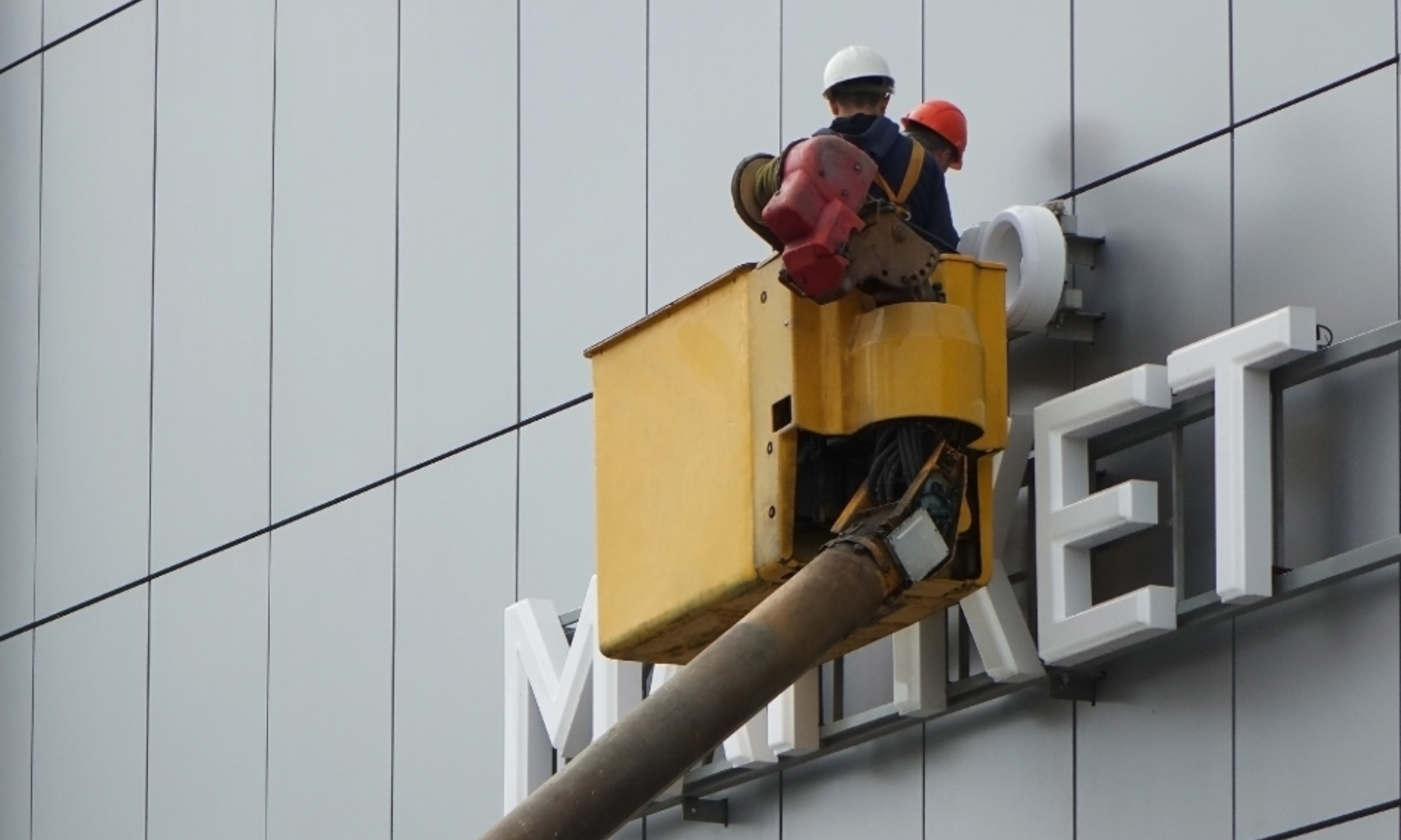 Two workers wearing safety helmets working on a building exterior, installing or repairing a sign on a gray wall.