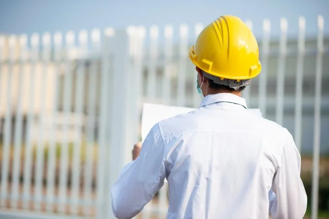 A person wearing a yellow safety helmet and white shirt, holding a clipboard, standing outdoors near a white fence.