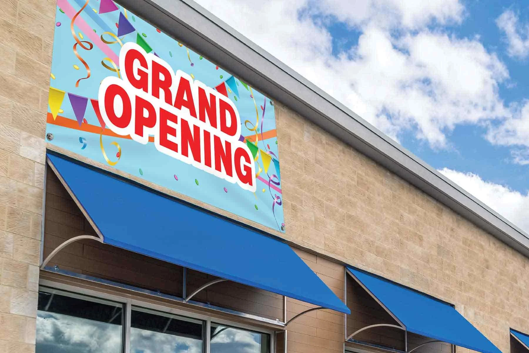 Colorful sign reading Grand Opening with blue awnings on a building against a partly cloudy sky.