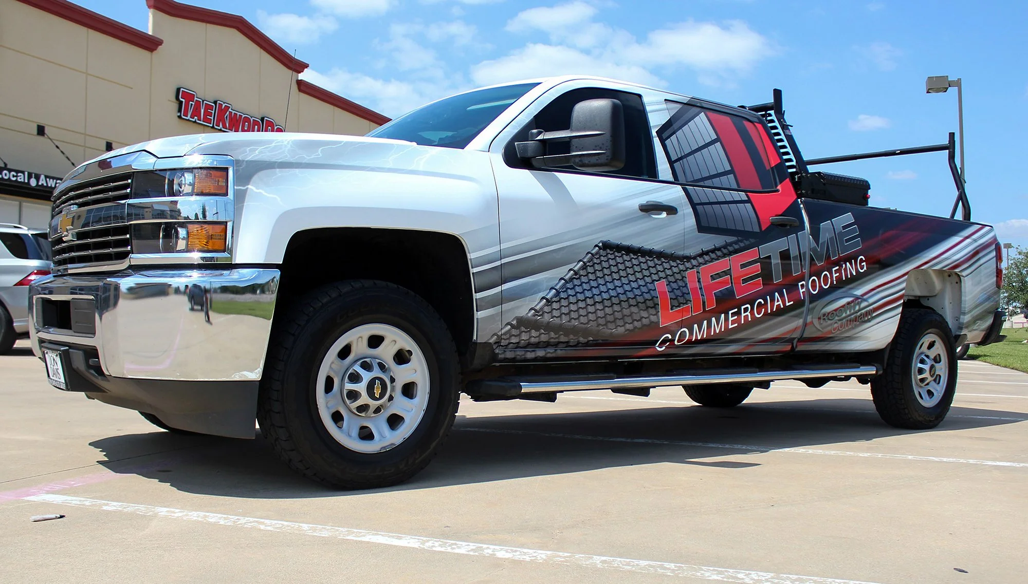 Commercial roofing company's pickup truck parked in a lot with building in background. The truck has branding for 'LIFETIME COMMERCIAL ROOFING'.