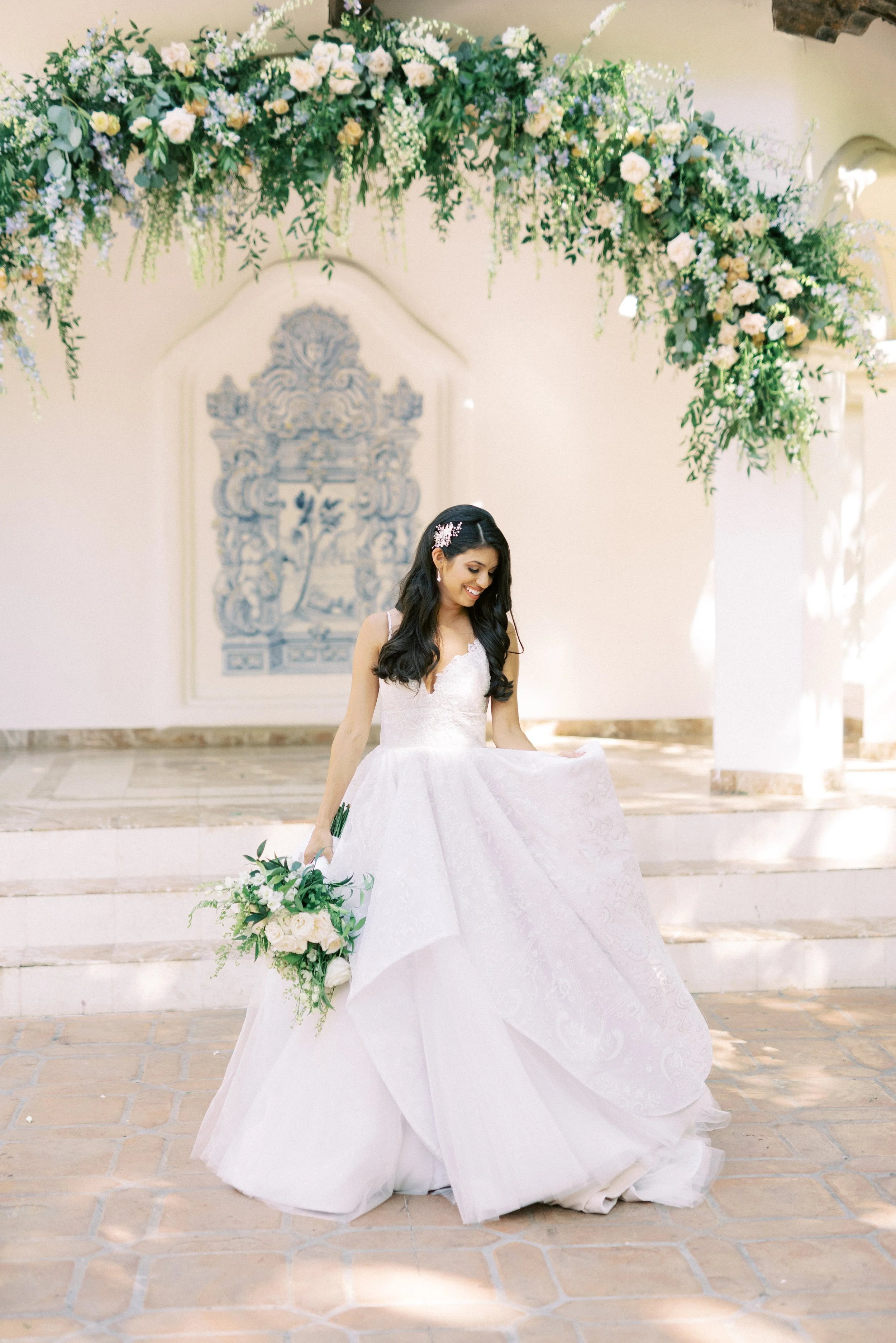 Bride in a white wedding dress holding a bouquet, standing under a floral archway.