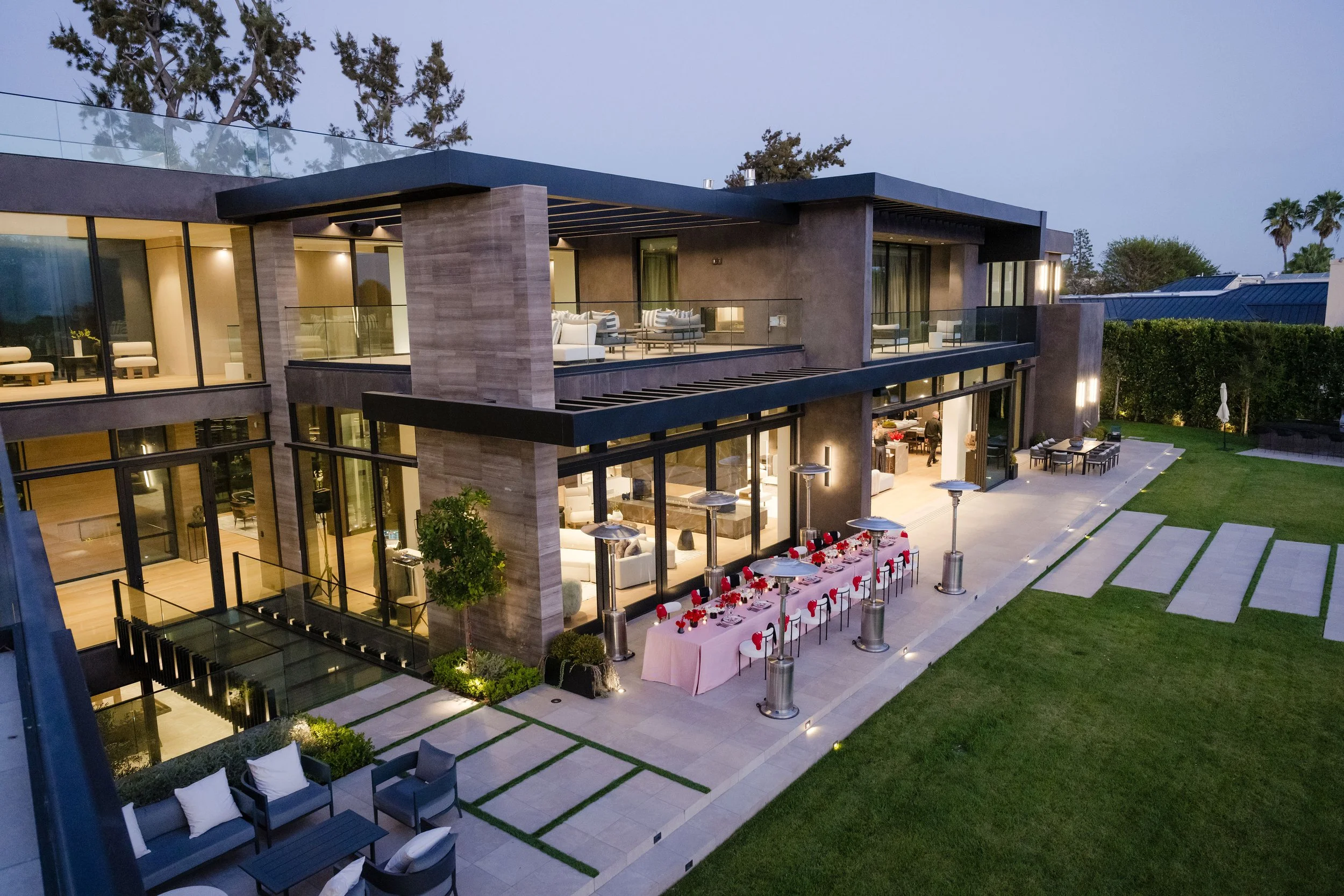 Modern luxury house exterior with large glass windows, outdoor patio with dining table setup, and well-maintained lawn at dusk.