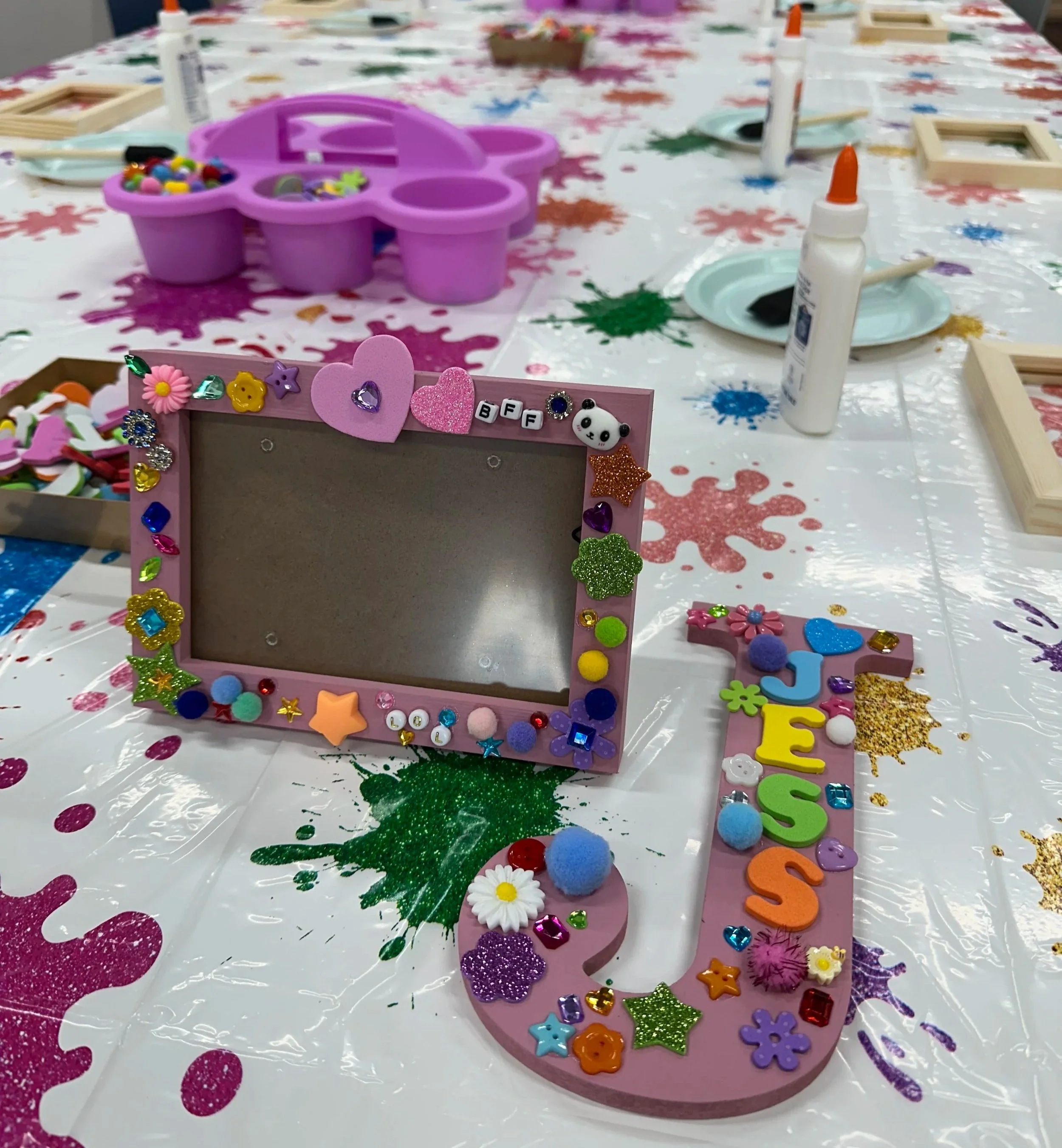 Colorful letter decorations spelling out "YEN" adorned with stickers, pom-poms, and small embellishments, surrounded by scattered glitter, paper charms, and decorations on a brown background.