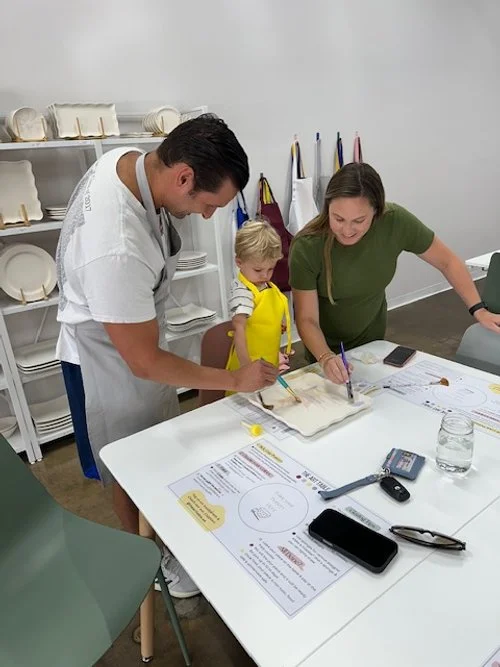 A person is painting orange paint onto a white ceramic bowl using a fine brush, holding it over a round, flat surface.
