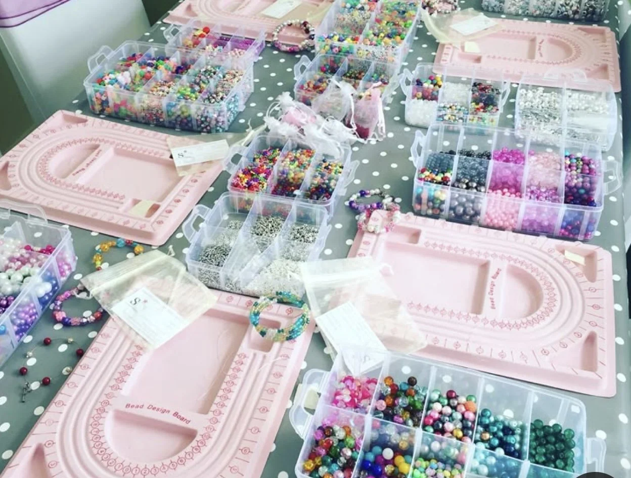 Table with multiple pink bead design boards and clear plastic containers filled with colorful beads and jewelry-making supplies.