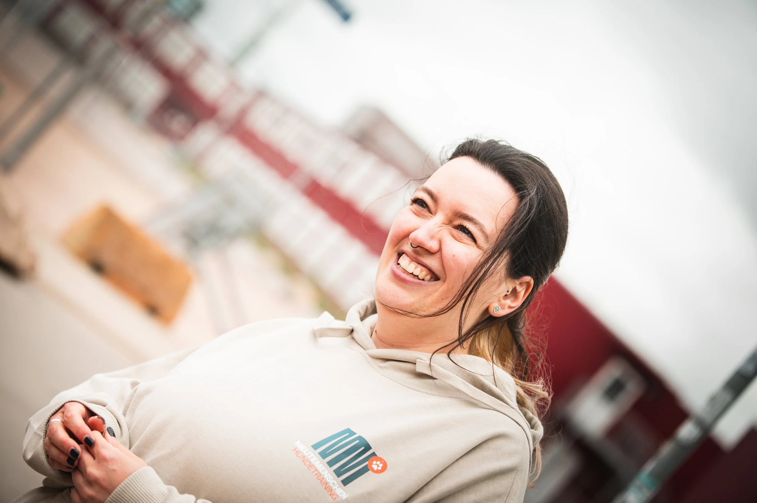 Lächelnde Frau mit dunklem Haar, trägt ein hellbeiges Hoodie mit Meutemenschen Logo. Sie ist locker gekleidet und schaut nach oben, während sie entspannt wirkt.