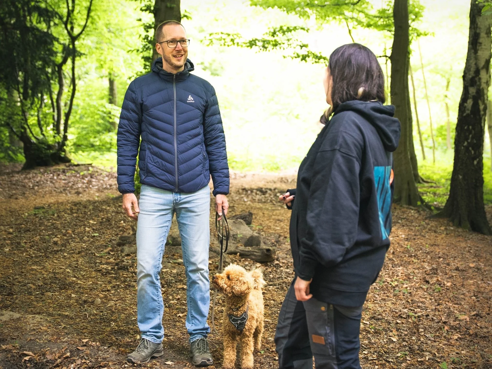 Zwei Menschen führen ein Hundetraining in einem Wald durch, zwischen ihnen ist ein Hund an der Leine.