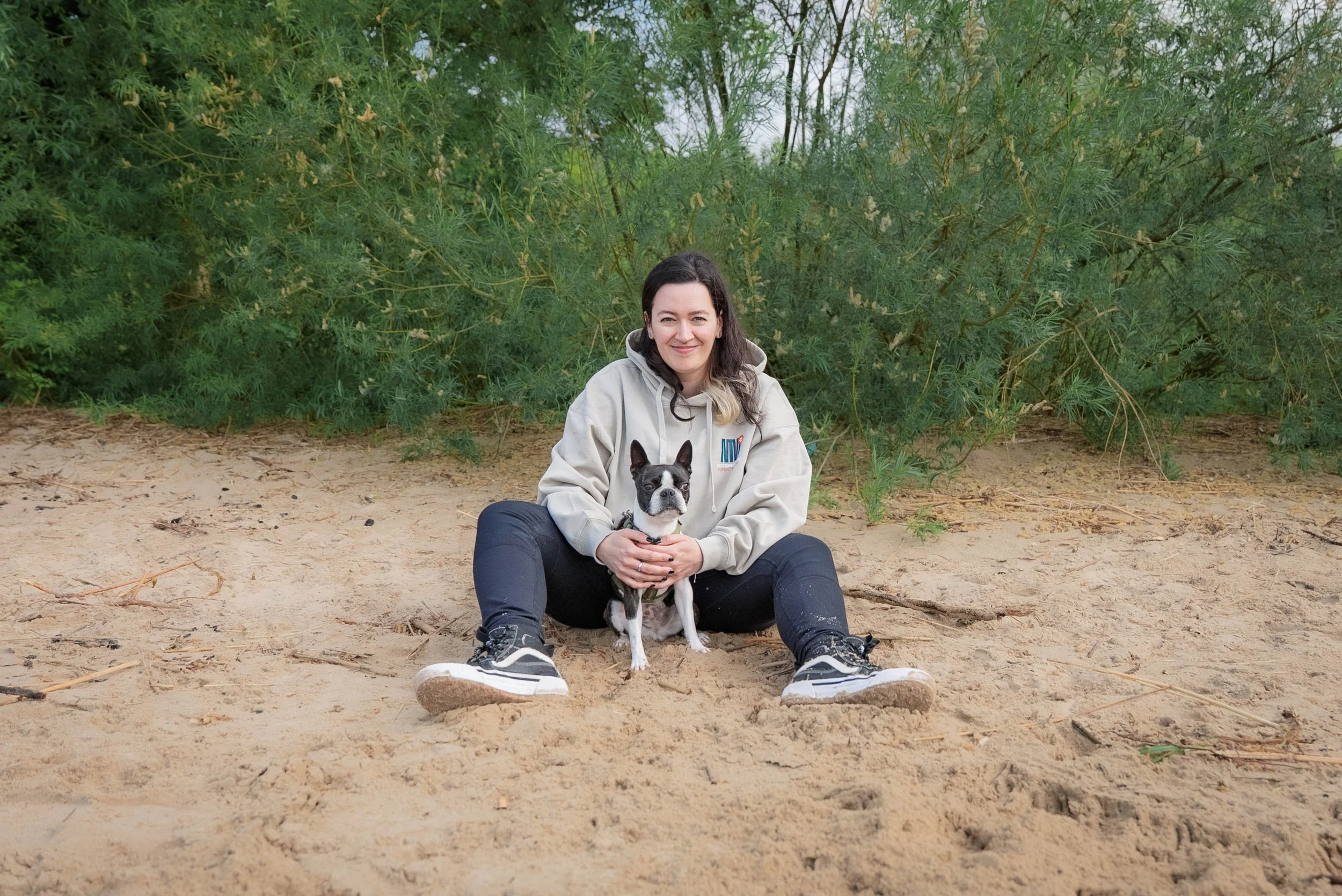 Eine Frau sitzt auf einem Sandstrand mit einem schwarz, weißen Hund, im Hintergrund dichte grüne Büschen.