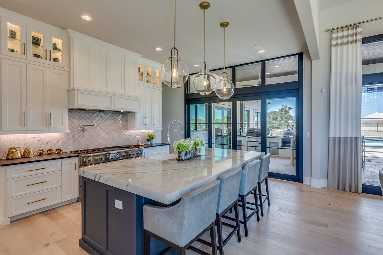 Modern kitchen with white cabinets, a large island with a light-colored marble countertop, pendant lighting fixtures, and glass doors leading outside with a view of an outdoor patio and BBQ grill.