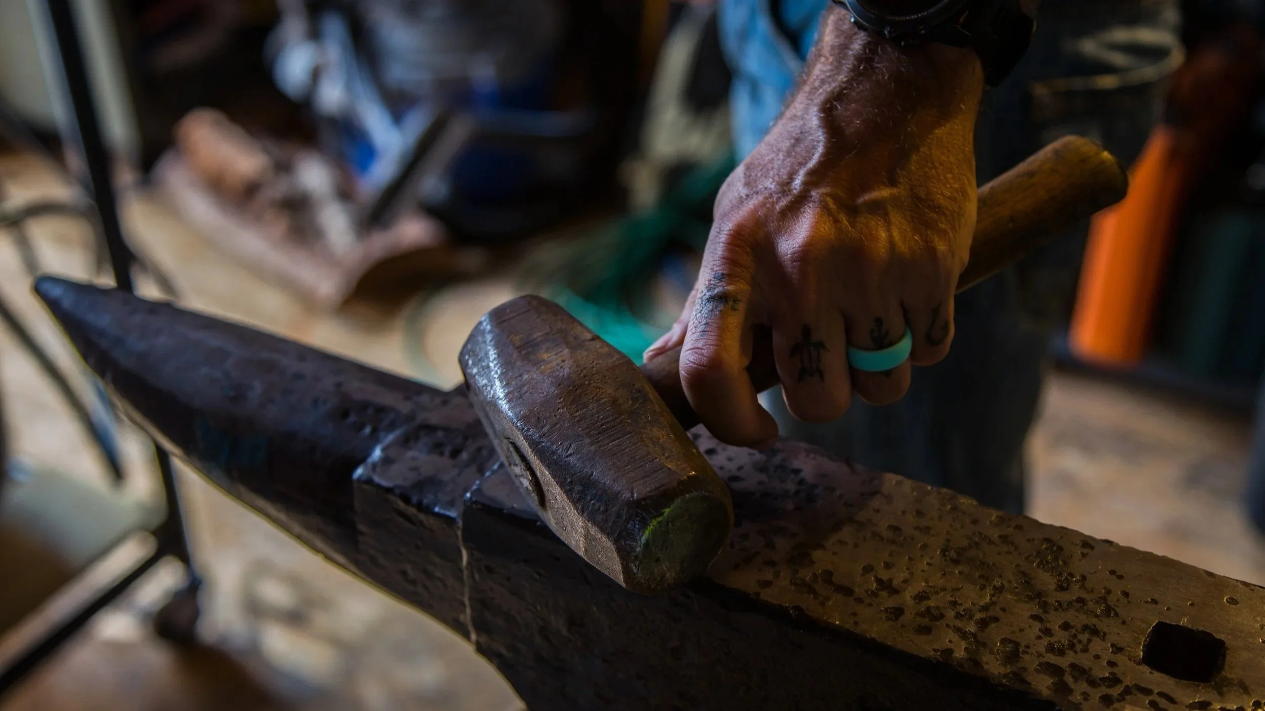 A person's hand holding a hammer, working on a piece of metal on an anvil in a workshop.