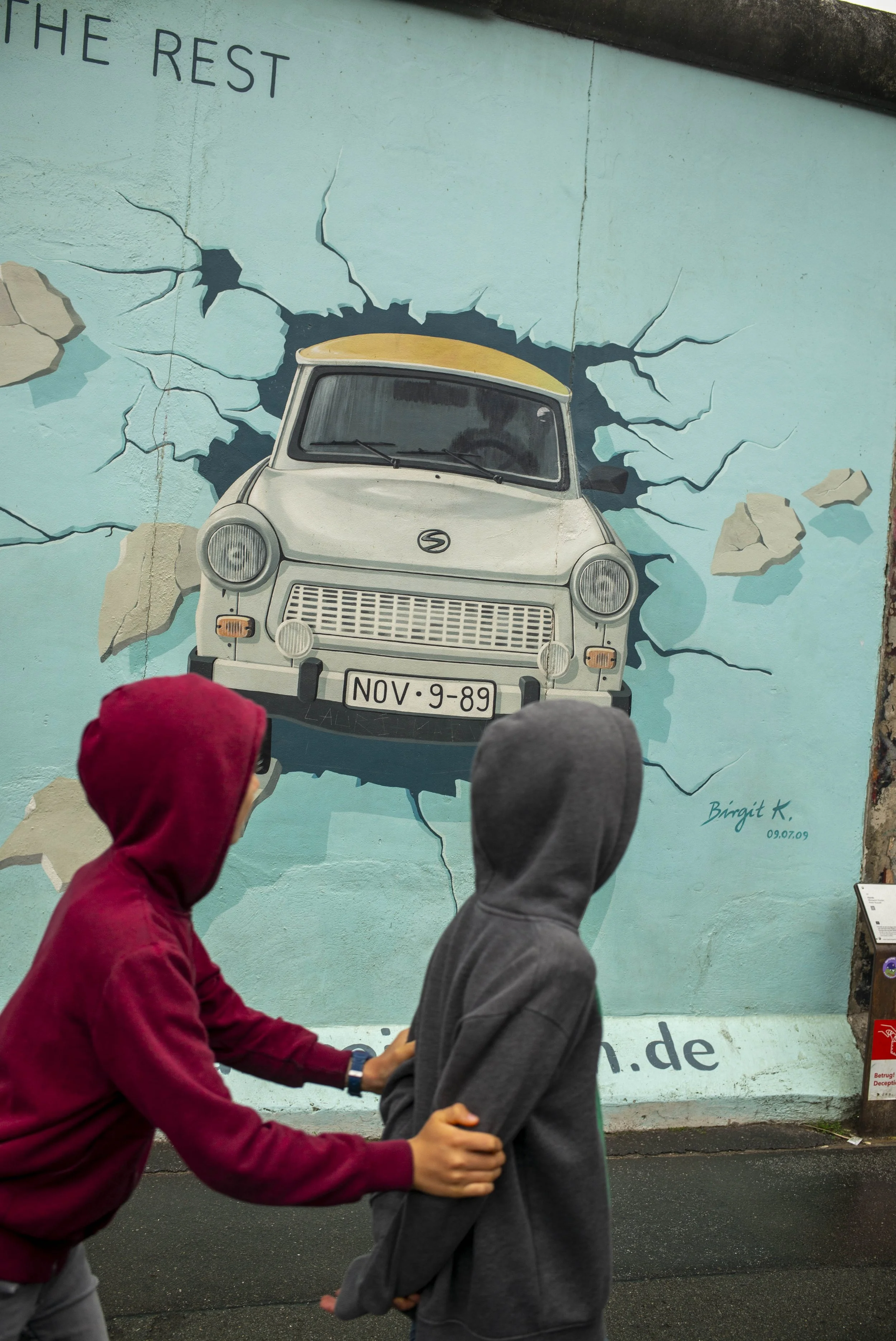 Un mural représentant une vieille voiture qui semble sortir d'un mur fissuré, avec deux enfants en hoodie passant devant.