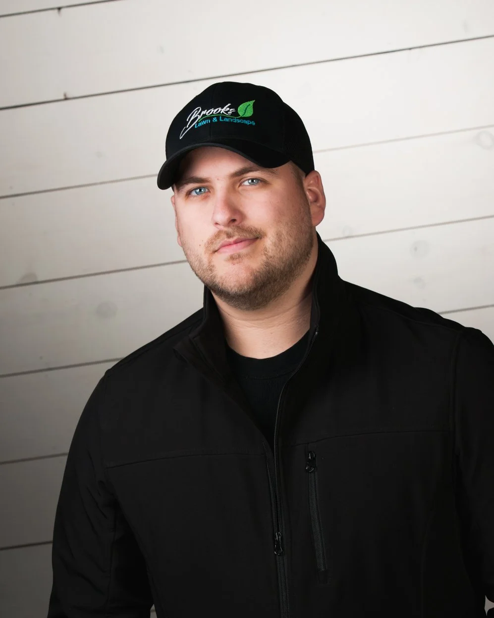 A man wearing a black cap with the logo 'Brooke Lawn & Landscape' and a black zip-up jacket, standing against a light-colored wooden wall.
