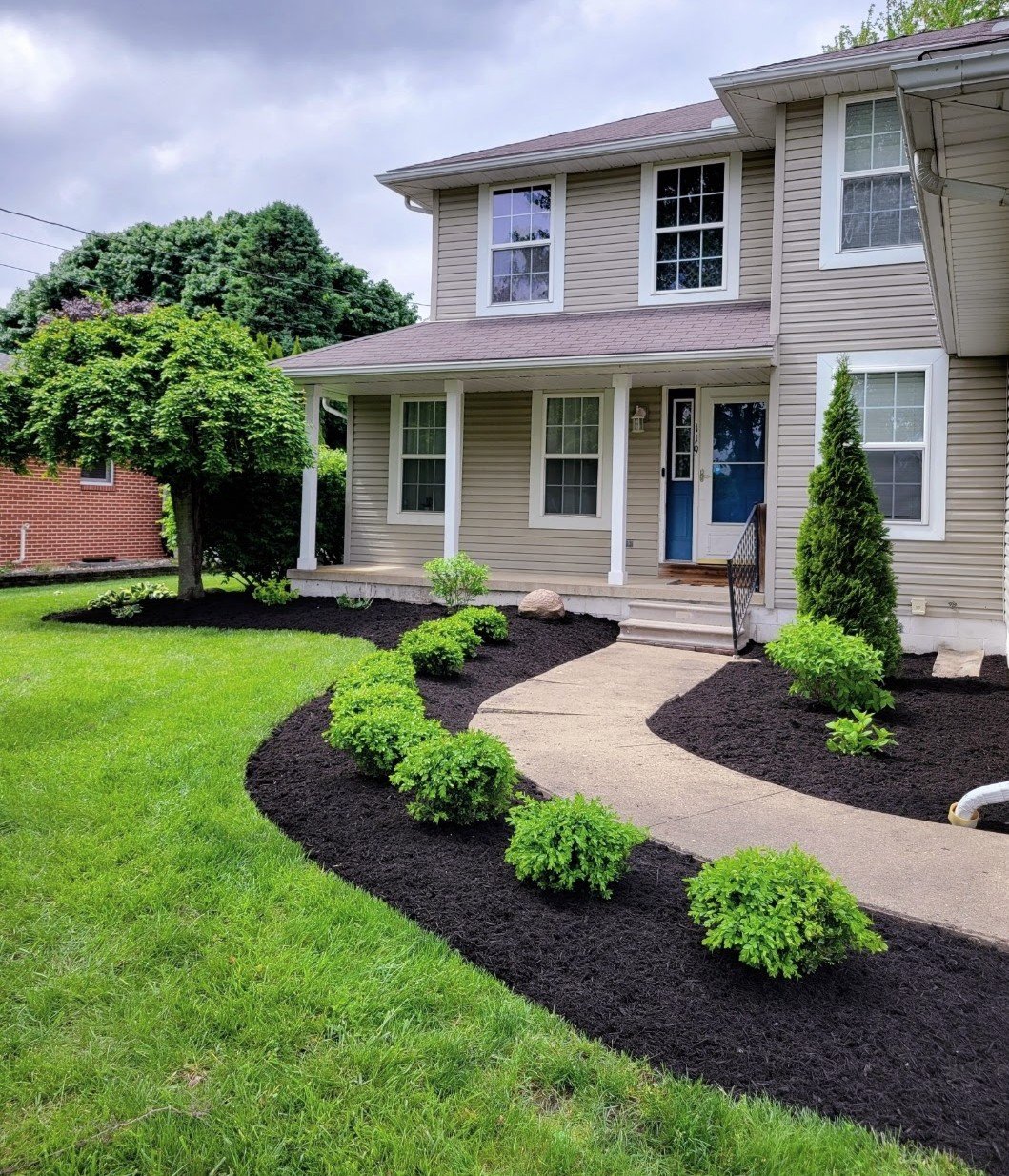 Front yard landscaped with green bushes along a curved walkway leading to a beige two-story house with white trim and a blue front door.