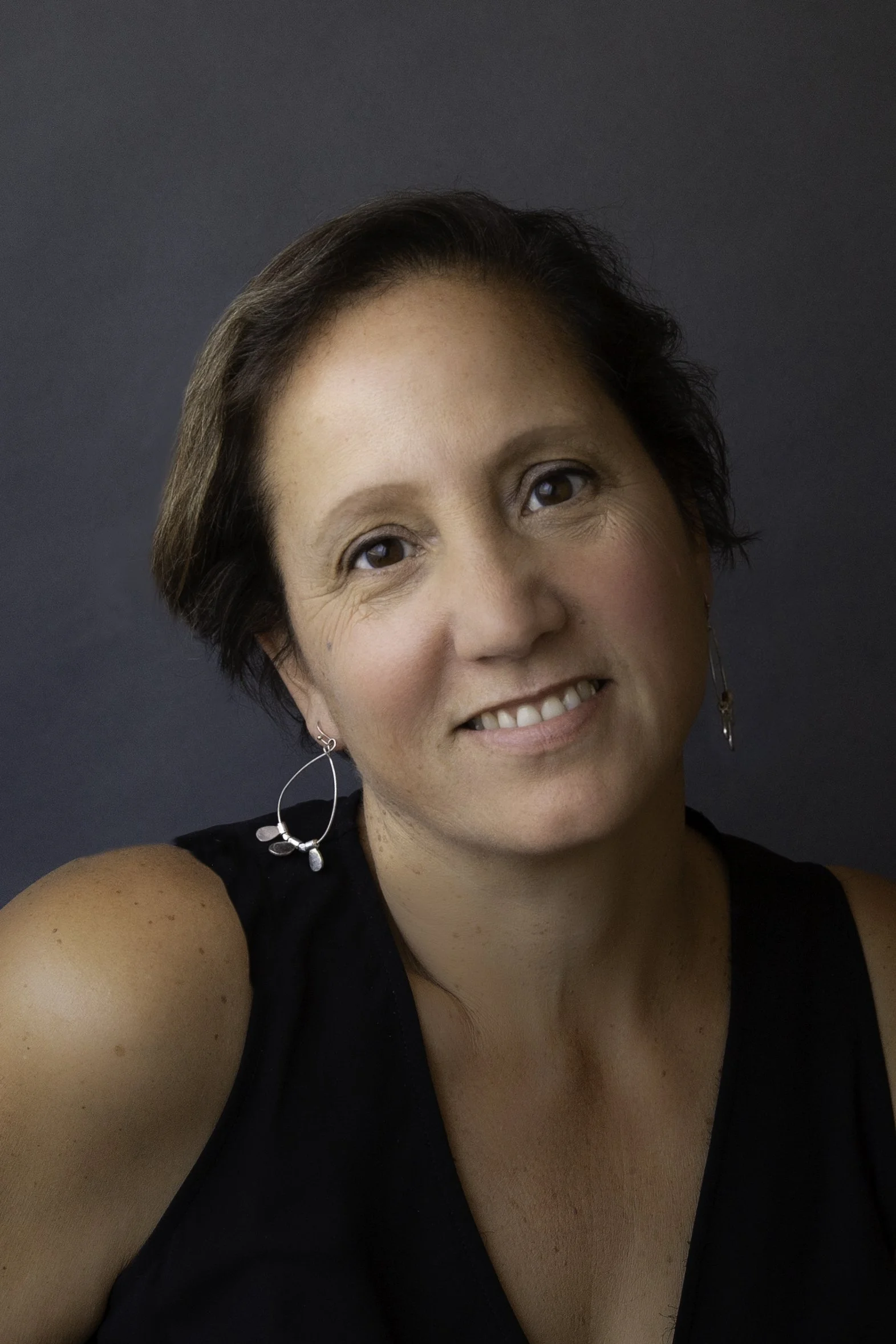 Close-up portrait of a woman with short brown hair, smiling, wearing a black sleeveless top and silver earrings, against a dark gray background.