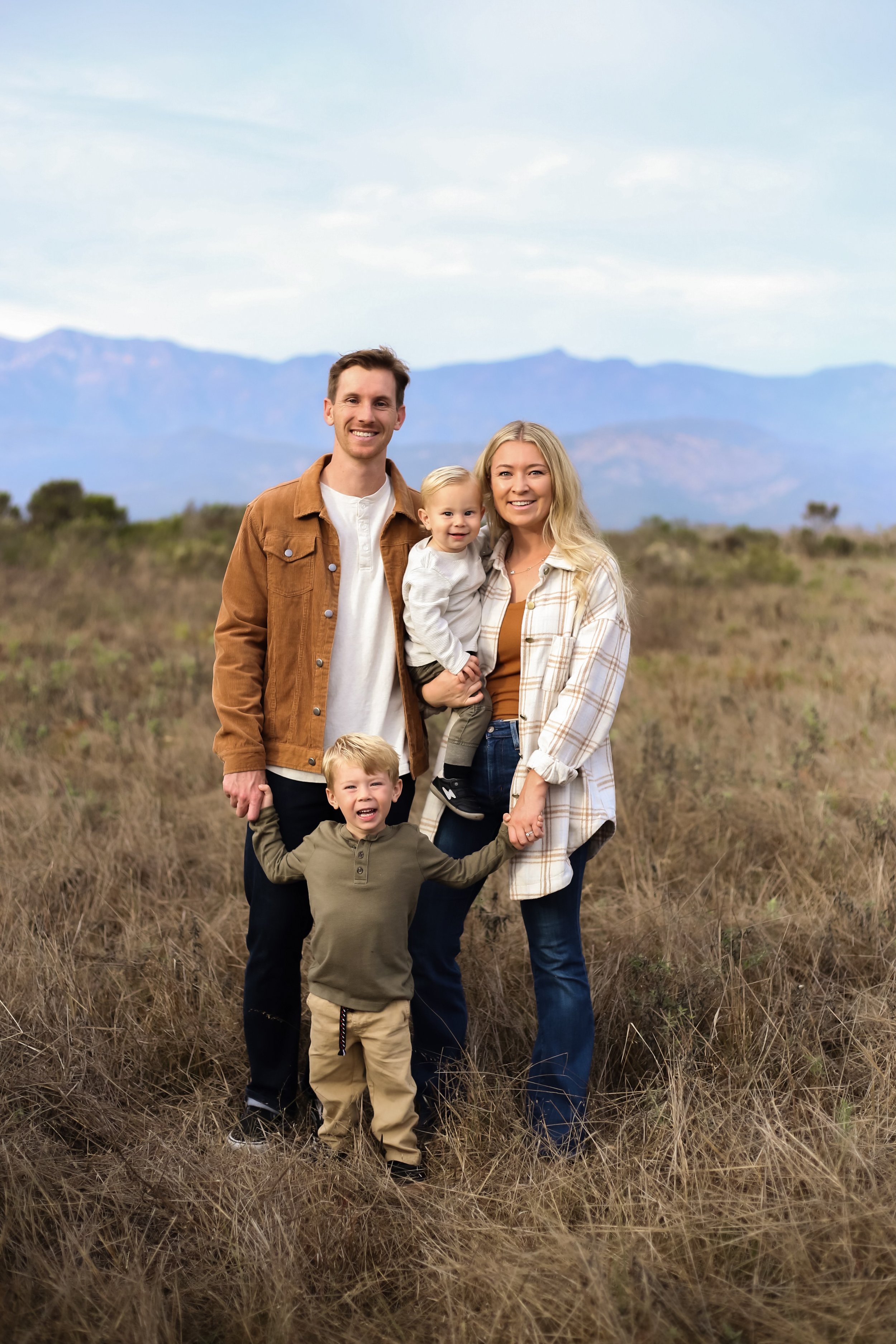 A family of four smiling and holding hands outdoors in a field with mountains in the background.