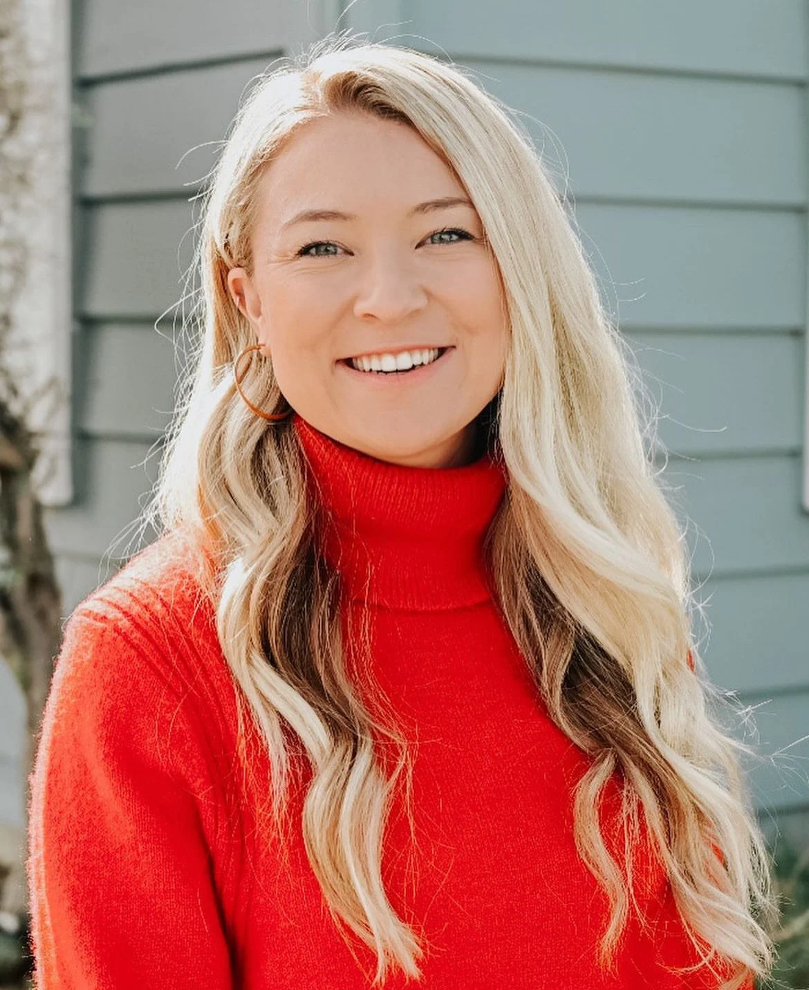 A woman with long blonde hair, wearing a red turtleneck sweater, smiling outdoors in front of a light gray house with horizontal siding.
