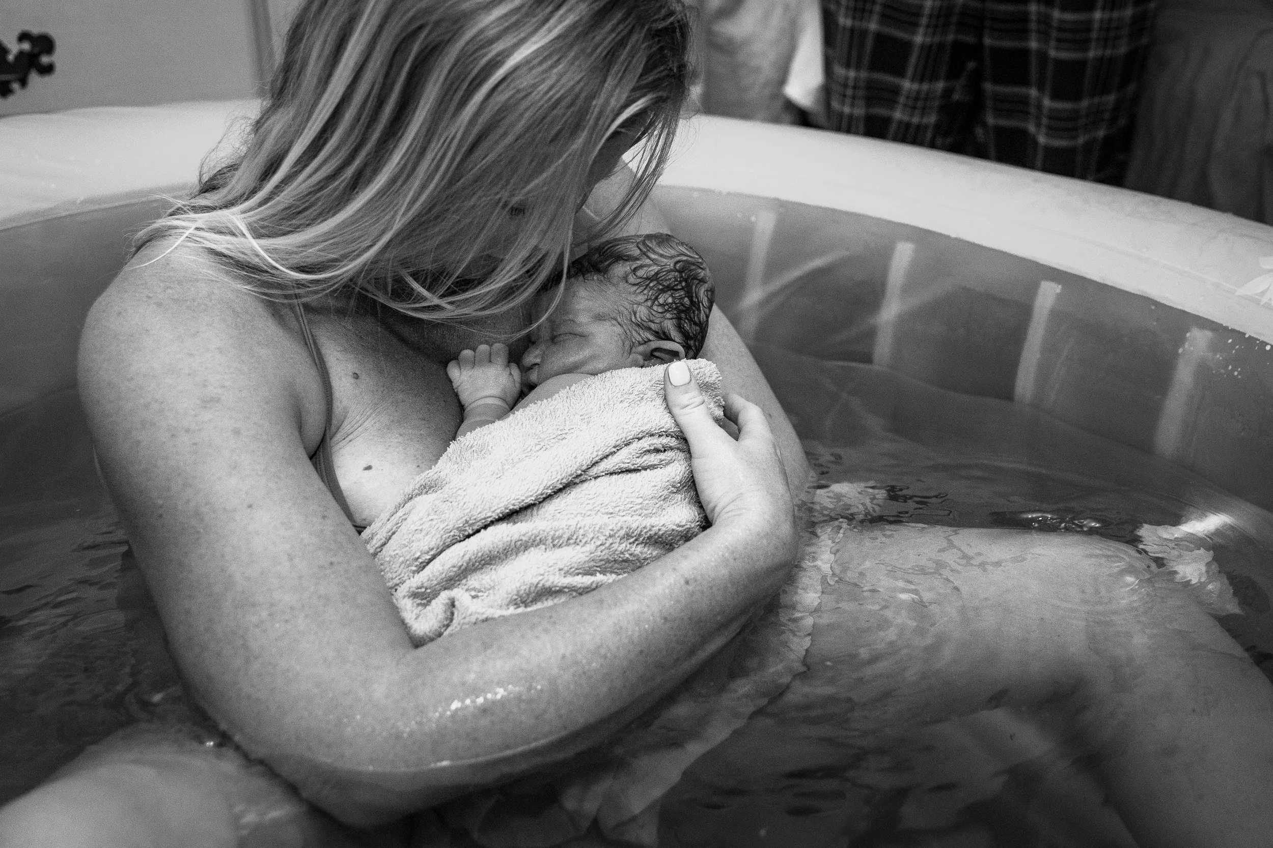 A woman holding a newborn baby wrapped in a towel during a water birth in a hospital or medical setting.