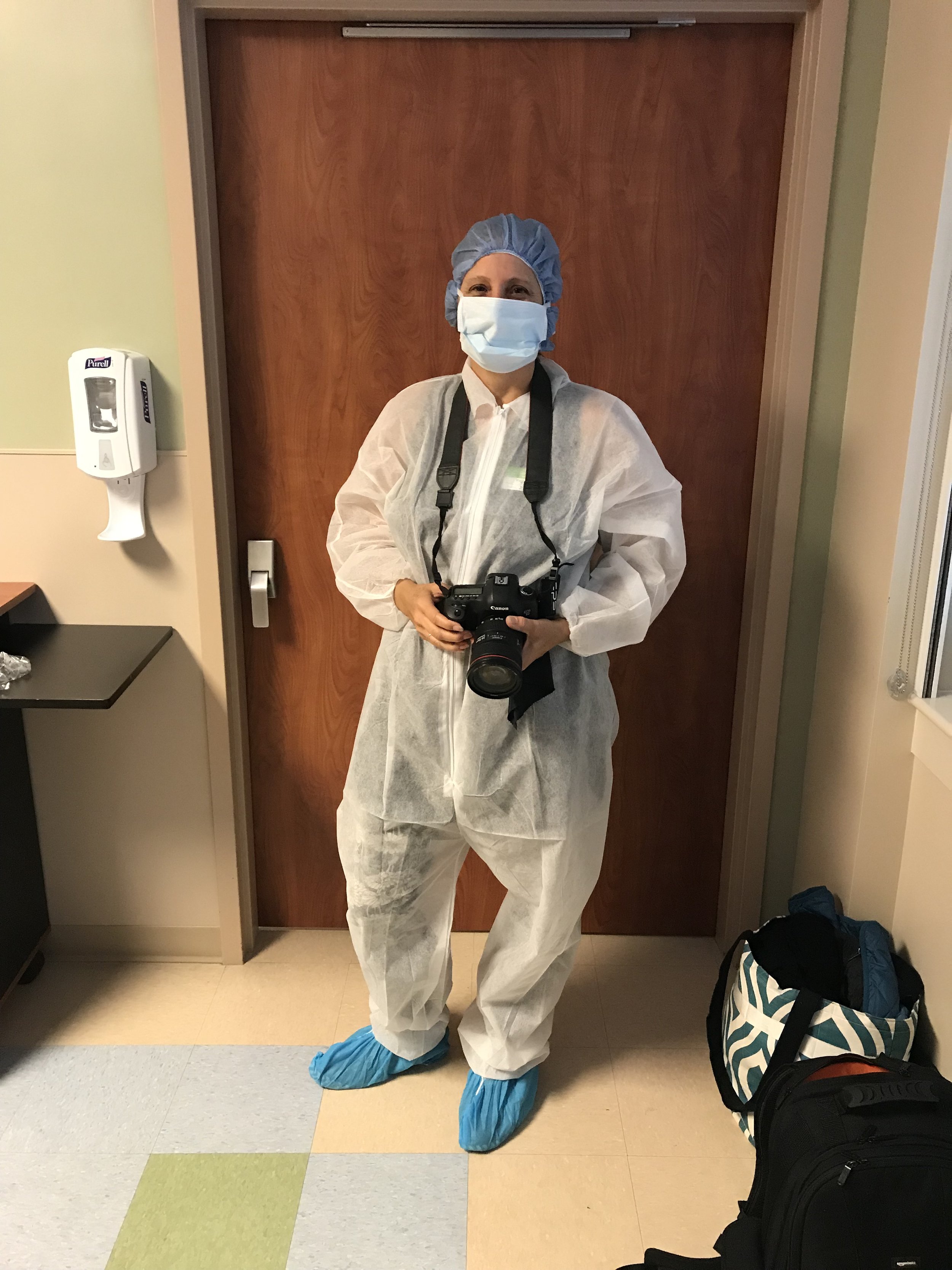A healthcare worker in full protective gear standing in front of a wooden door in a hospital. She is wearing a blue surgical cap, a face mask, a white protective gown, blue shoe covers, and holding a camera.