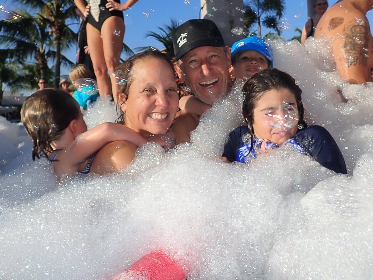 Group of people smiling and laughing in a foam-filled hot tub outdoors with palm trees in the background, enjoying a sunny day.