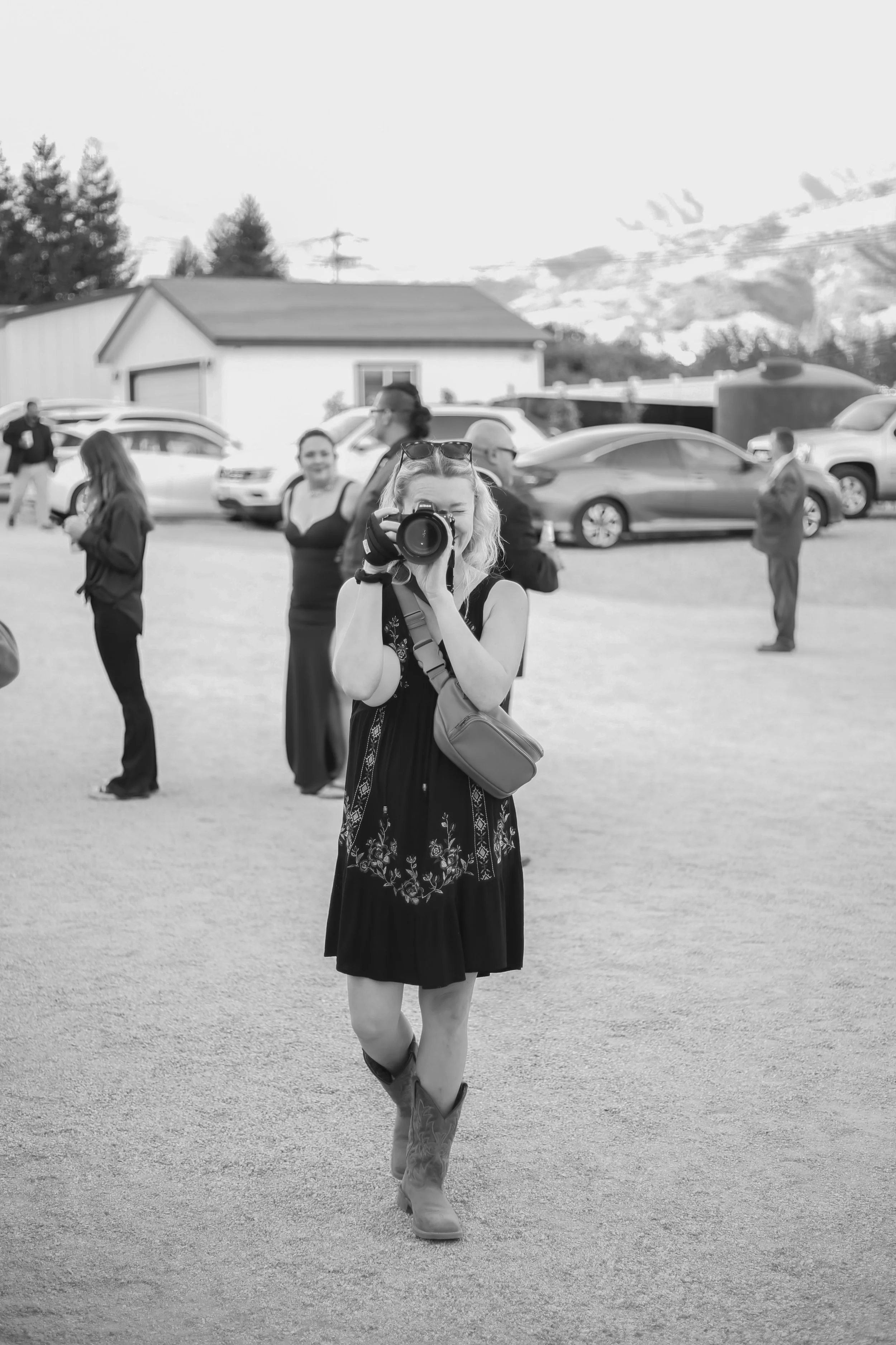 A woman taking a photo with a camera outdoors, wearing a dress and cowboy boots, standing in front of a group of people, with cars and a building in the background.