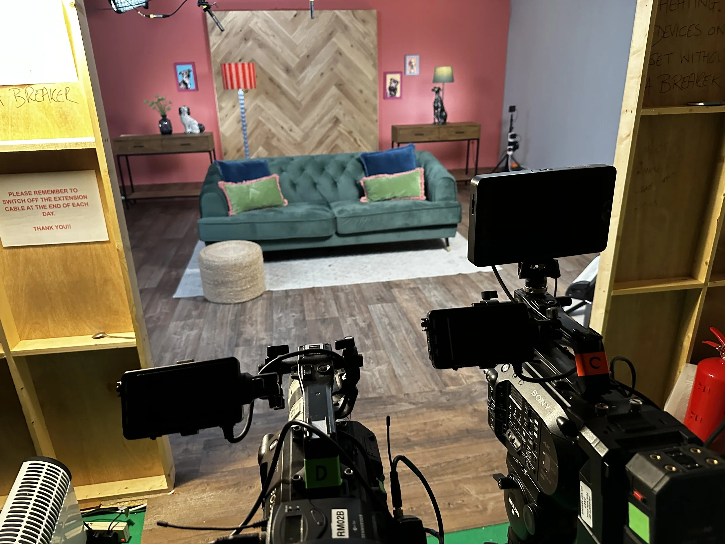 A behind-the-scenes view of a video shoot or filming setup for Tiger Ant Productions showing the camera equipment focused on a staged living room scene with a teal sofa, colorful pillows, and wall decor. Tigerantproductions