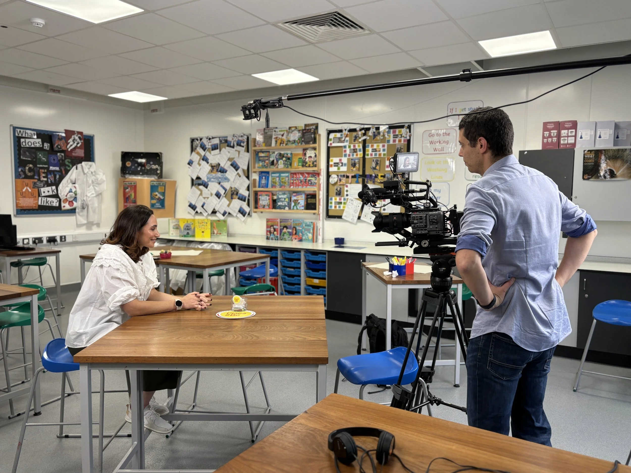 Tiger Ant Productions on set - A woman sitting at a school desk being filmed by a man with a professional camera in a classroom.
