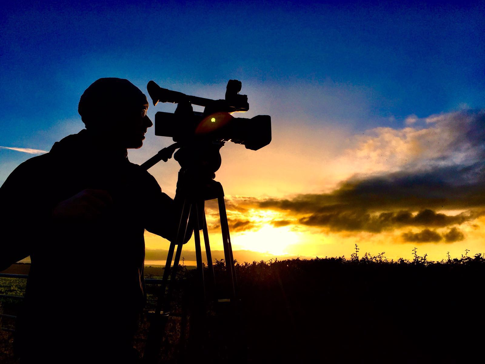 Silhouette of a person operating a video camera on a tripod during a sunset with colorful sky.