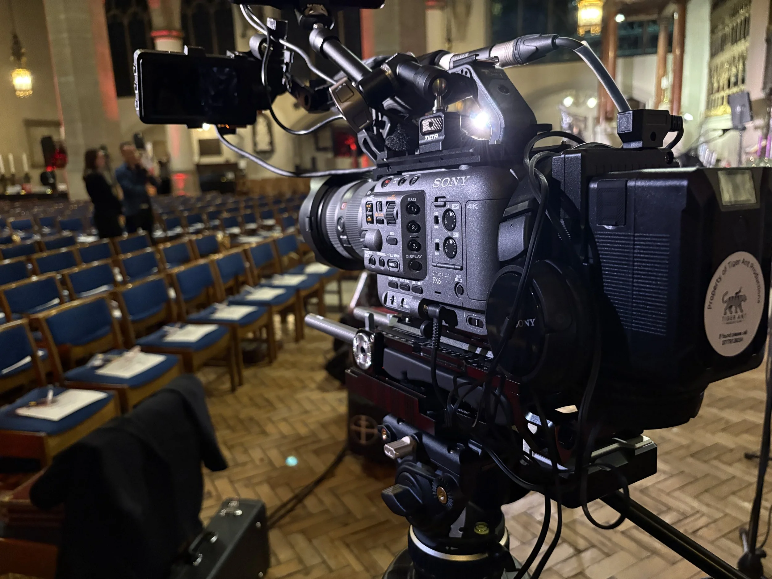 Tiger Ant Productions on set - A professional Sony video camera setup indoors at an event venue with empty chairs arranged in rows, some with napkins and utensils, and a few people in the background.