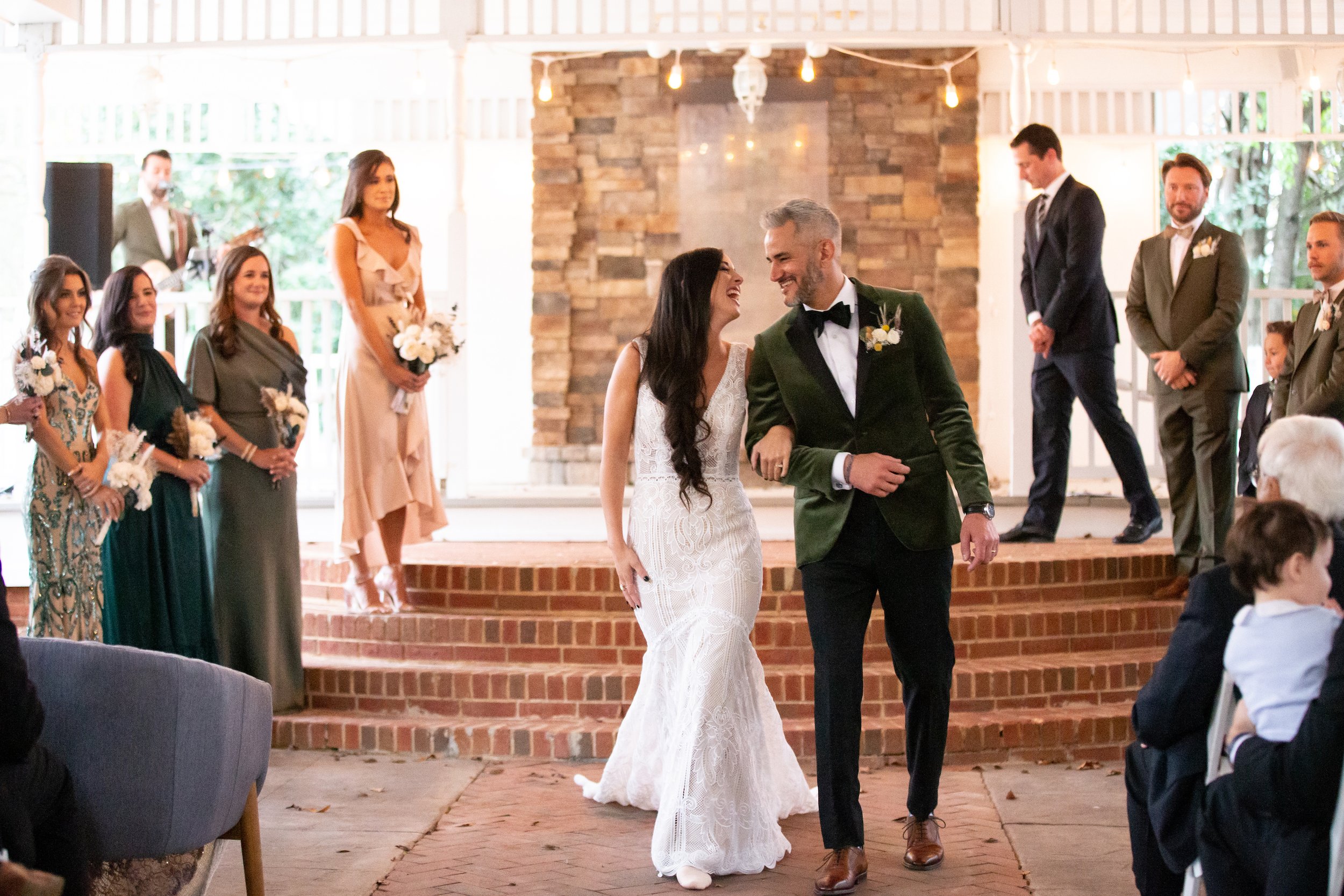 A bride and groom walking arm in arm during their wedding ceremony, surrounded by bridesmaids and groomsmen, with guests seated in the foreground.