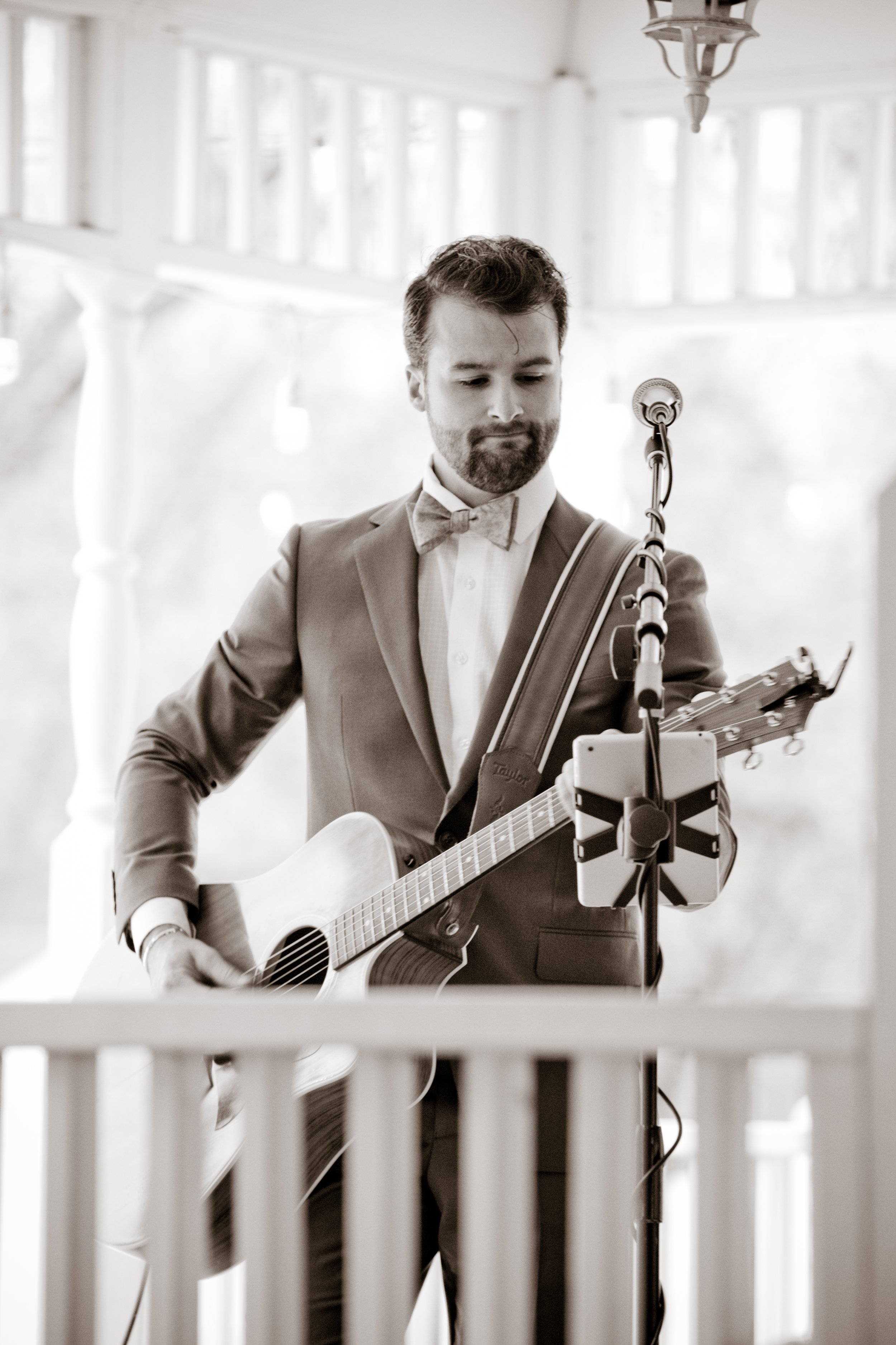 A man in a suit with a bow tie playing an acoustic guitar at a microphone stand in a decorated indoor setting.