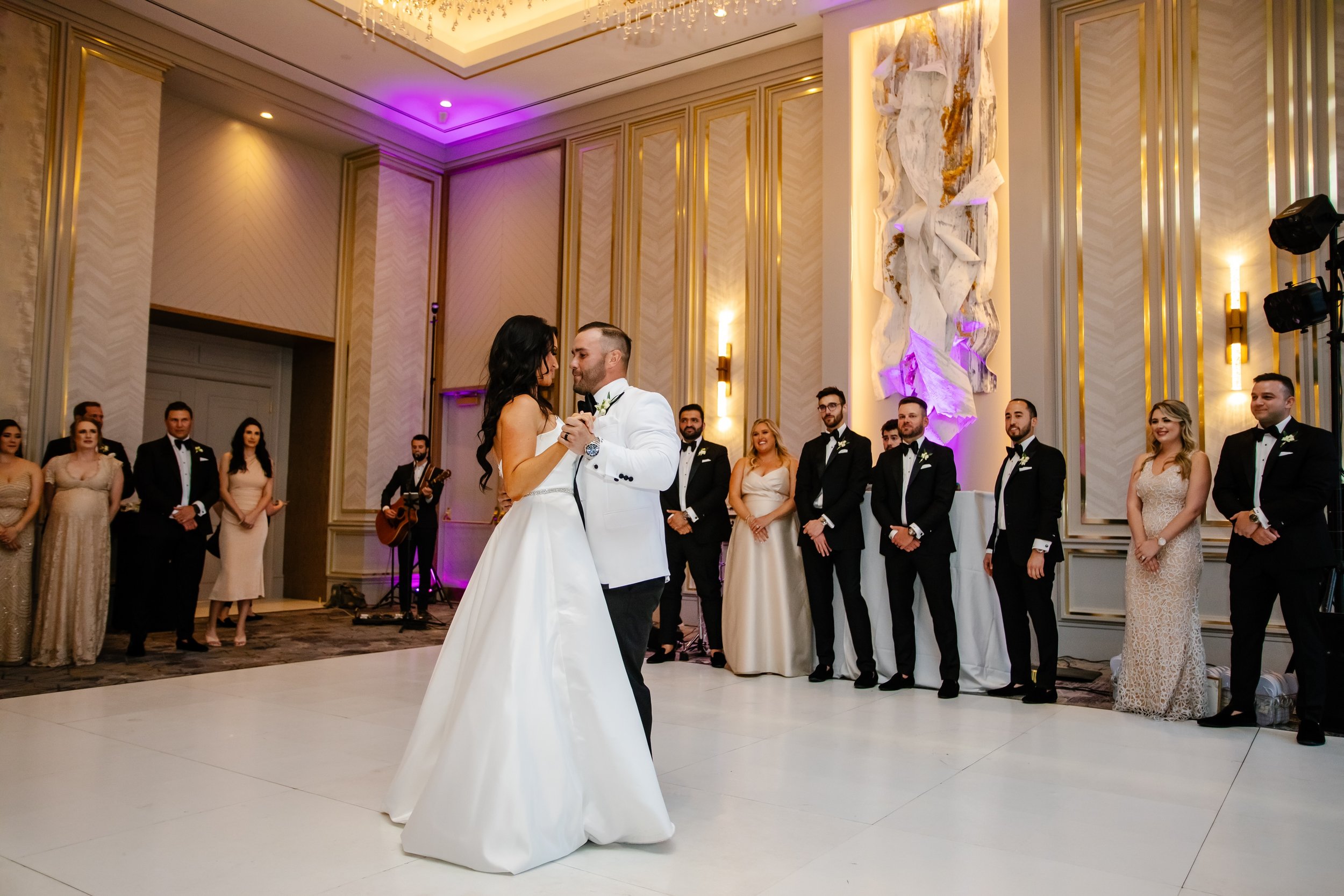 A bride and groom share a dance at their wedding reception with guests standing around them and a live guitarist singer playing in the background.