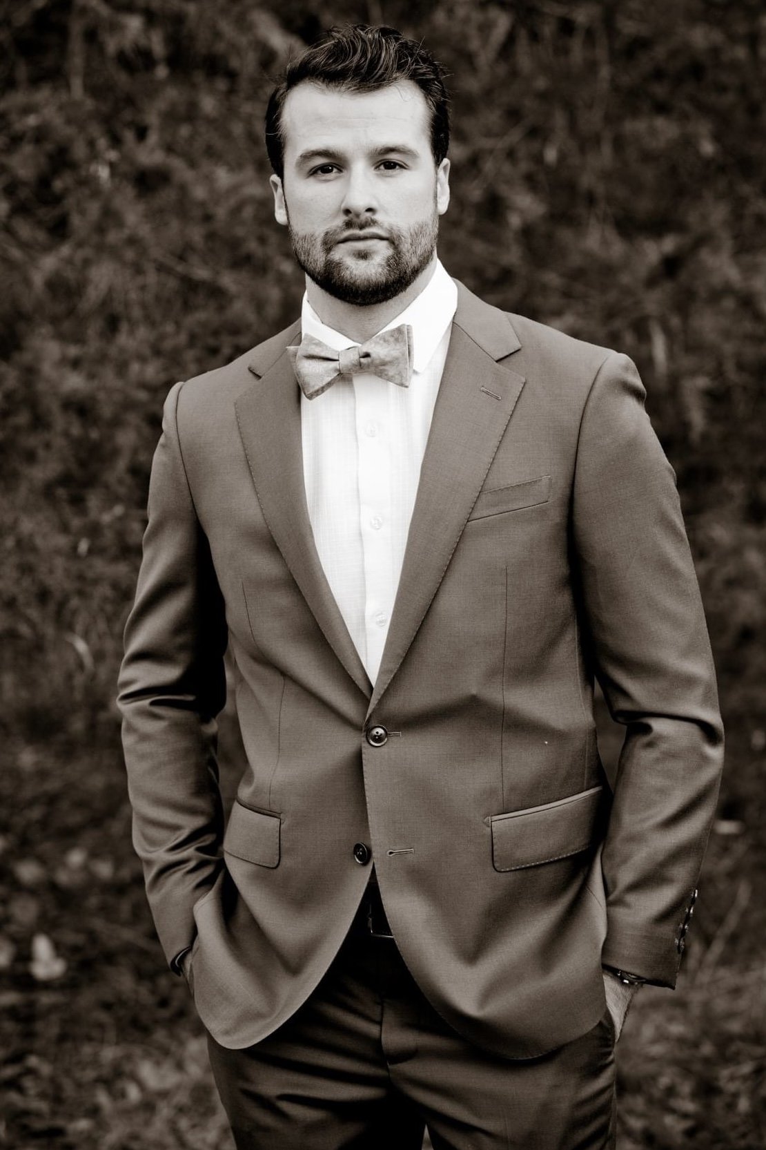 A man in a suit and bow tie standing outdoors, looking at the camera, in black and white.
