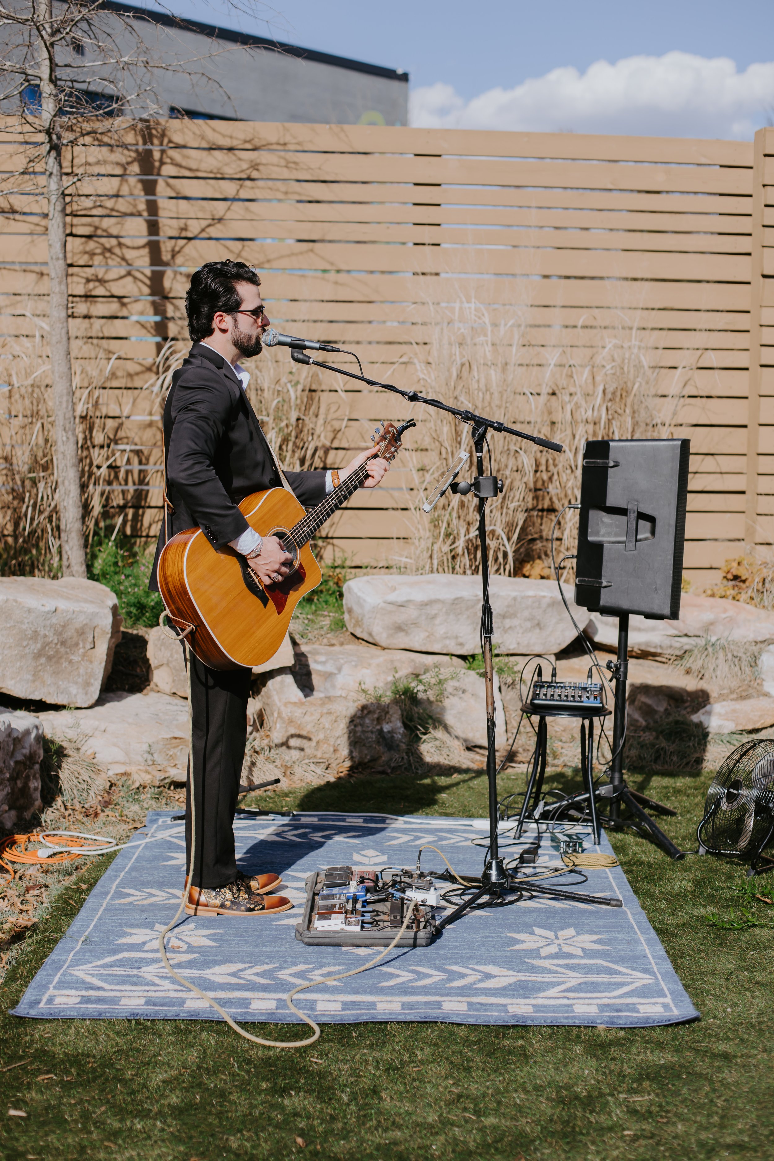 A man in a black suit and sunglasses playing acoustic guitar and singing into a microphone outdoors during the daytime. There is audio equipment, a speaker, and a small amplifier on a stand around him, with rocks, dried grass, and a wooden fence in t