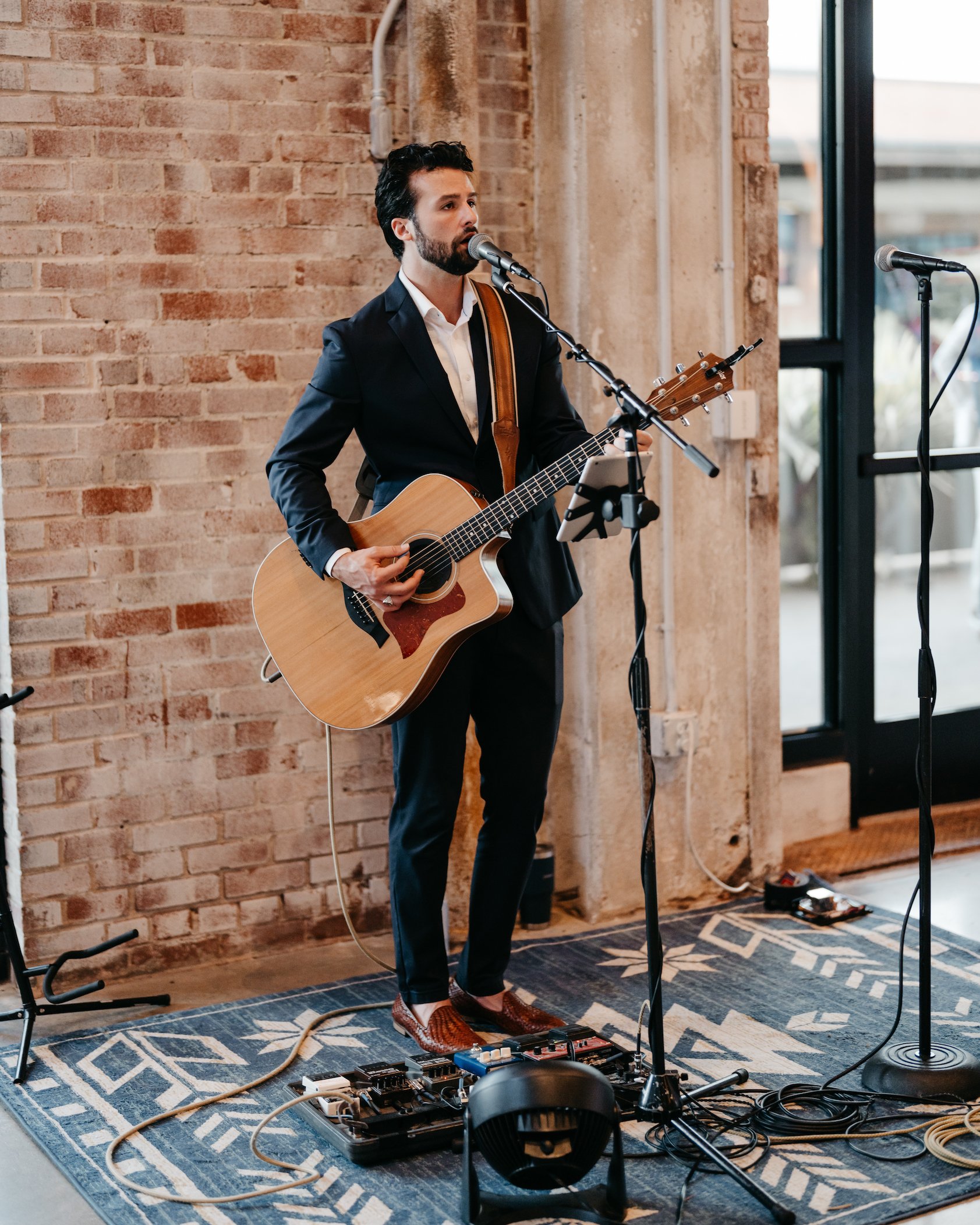 A man in a black suit playing an acoustic guitar and singing into a microphone indoors, with a brick wall and large windows in the background.