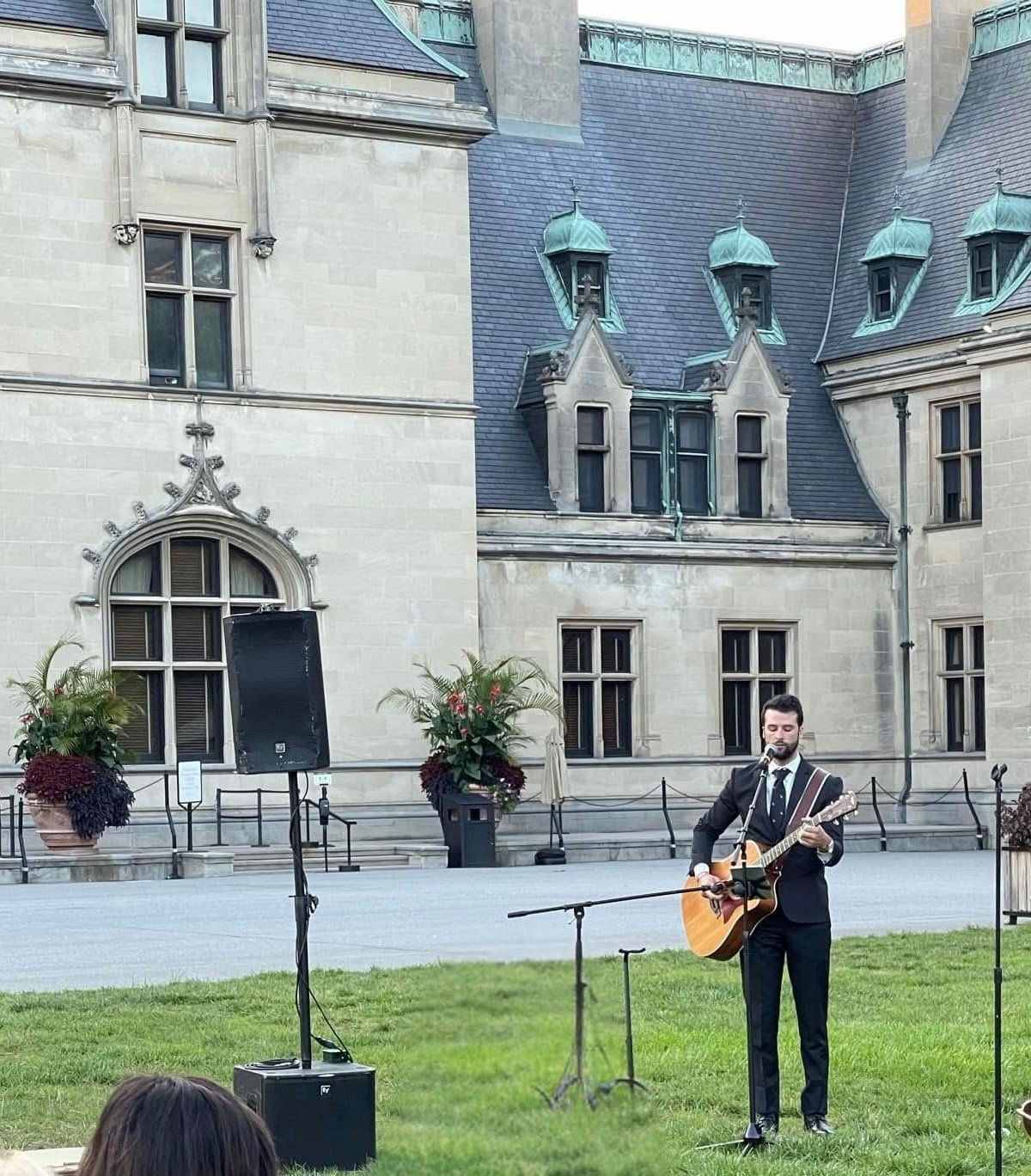 A man in a suit playing guitar and singing into a microphone outdoors, with a historic stone building in the background and potted plants nearby.