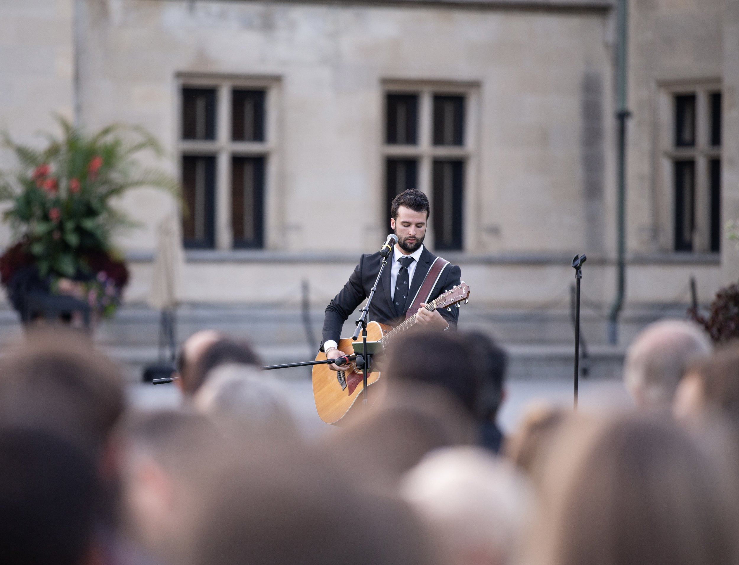 Man in a suit playing an acoustic guitar and singing into a microphone in front of an audience outdoors.