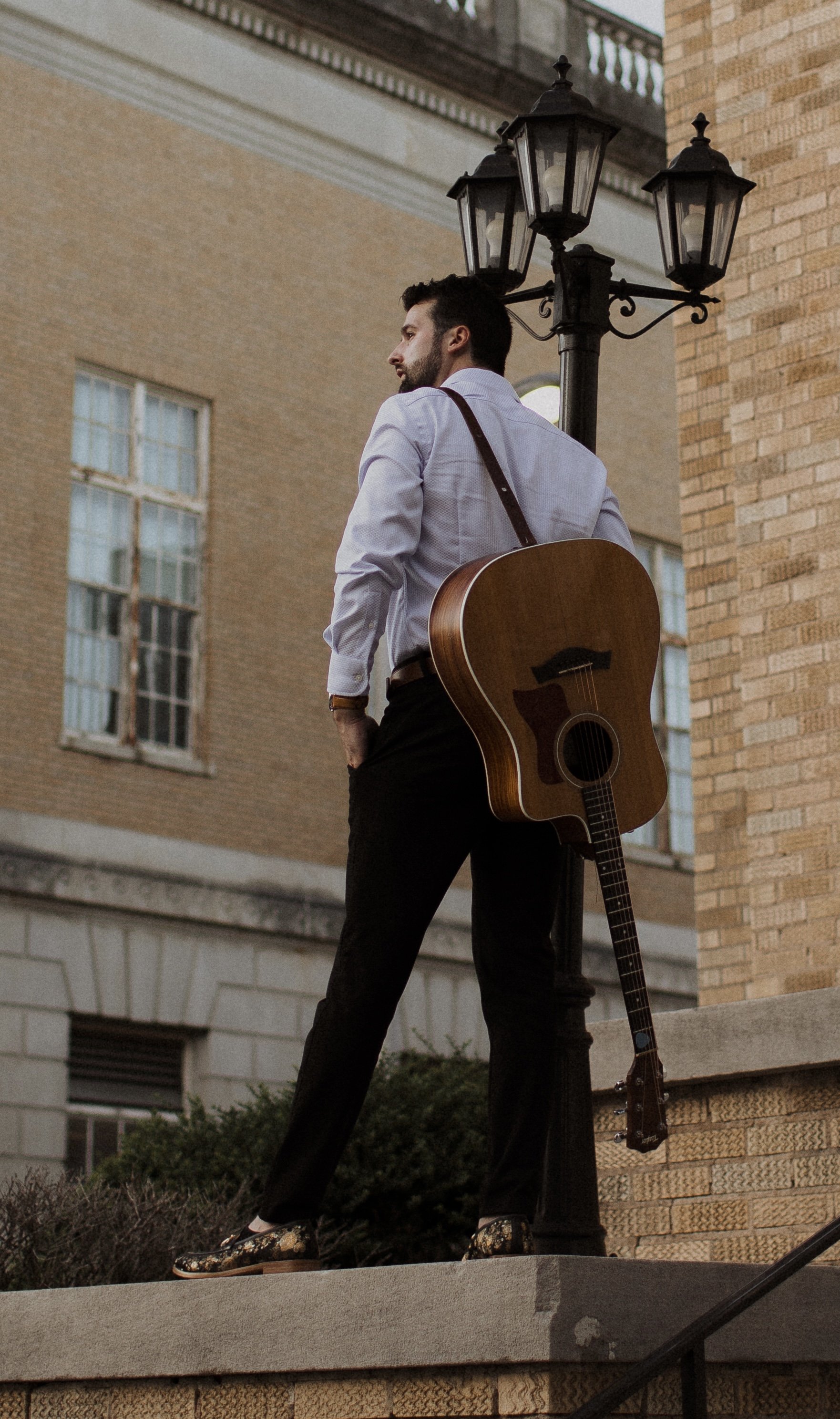A man standing outside near a brick building, leaning on a lamppost, with an acoustic guitar slung over his back, wearing a white shirt, dark pants, and floral shoes.