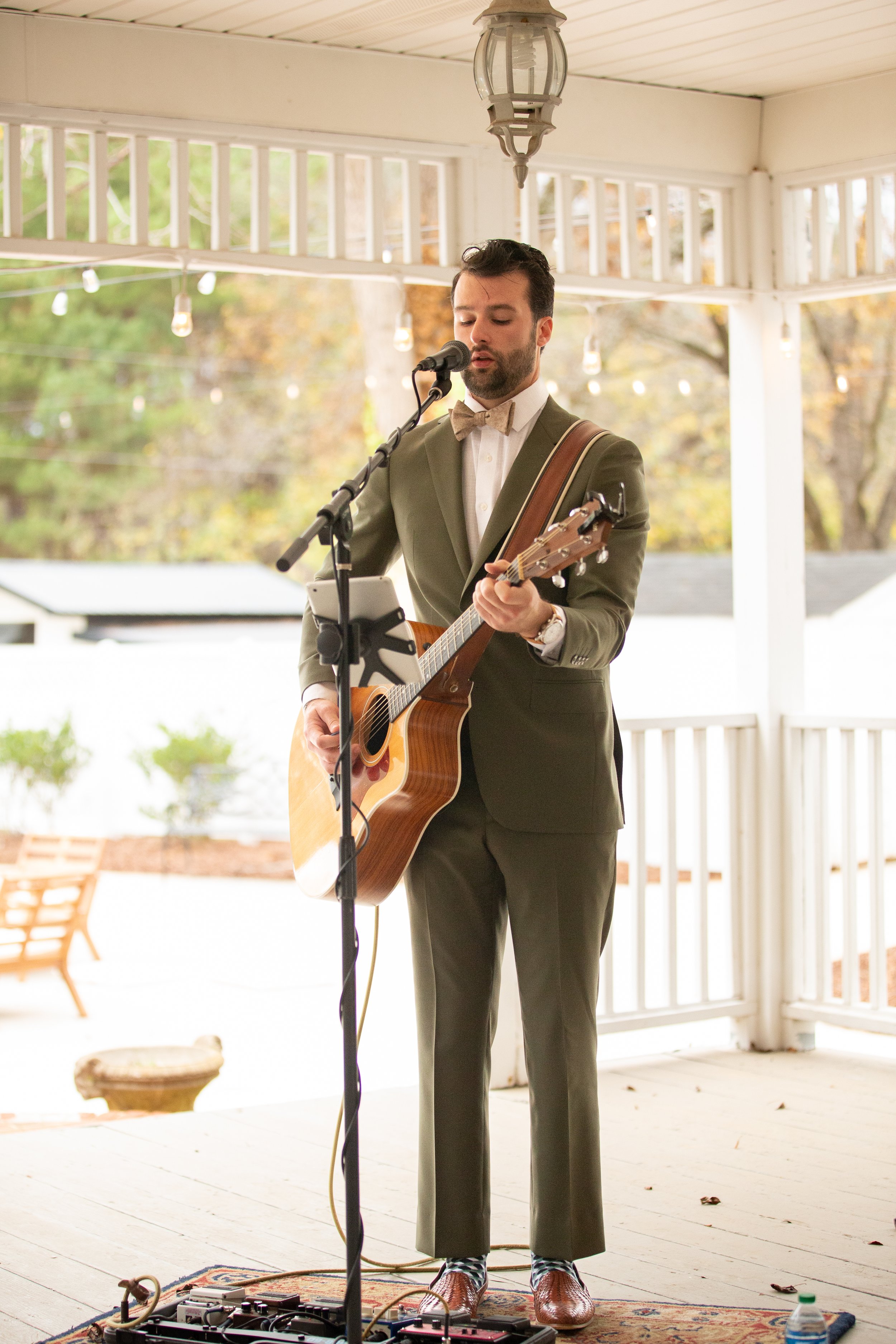 A man in a tuxedo with a bow tie and pants playing an acoustic guitar and singing into a microphone on a covered porch during daytime.
