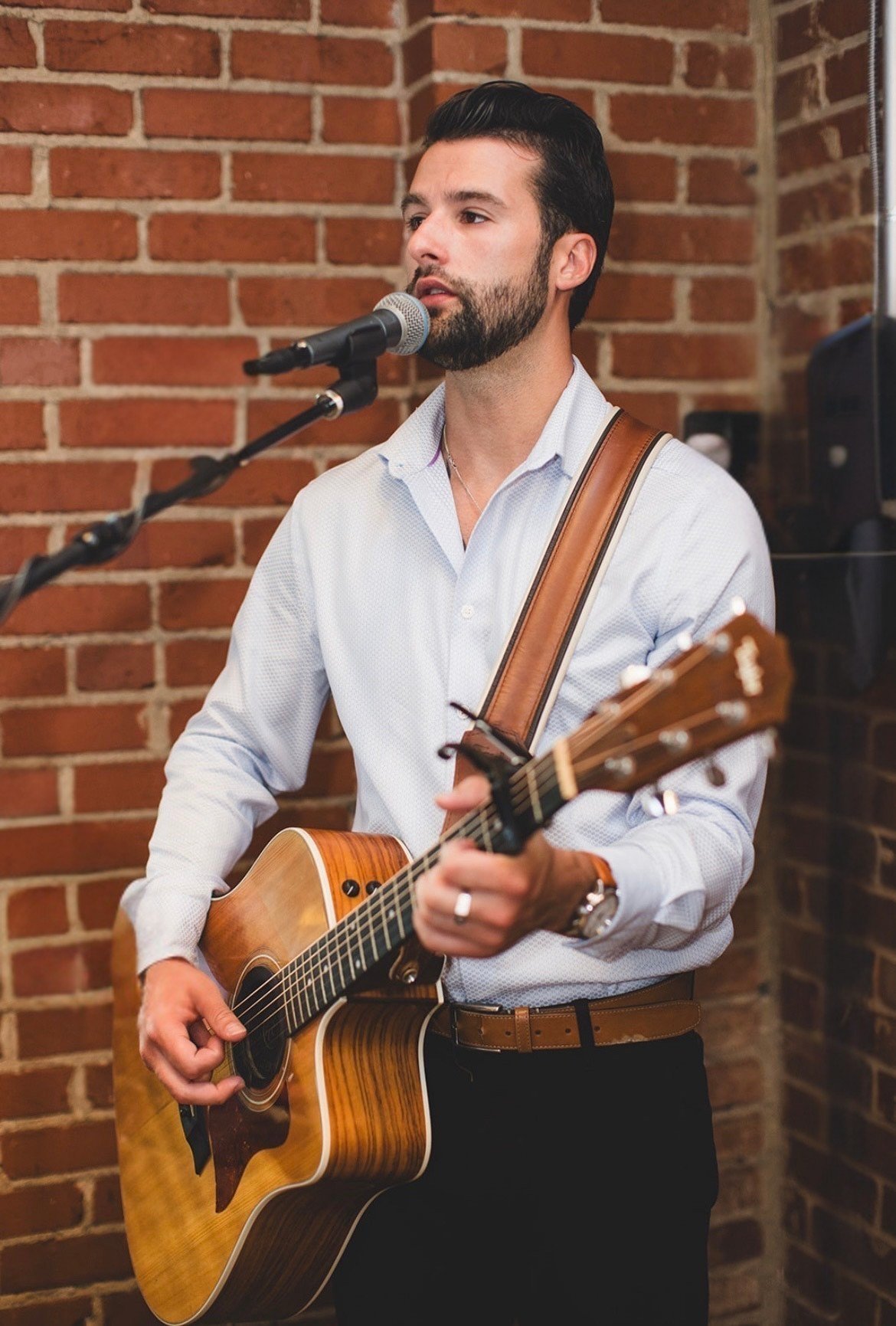 A man with dark hair and a beard playing an acoustic guitar and singing into a microphone, standing against a brick wall.