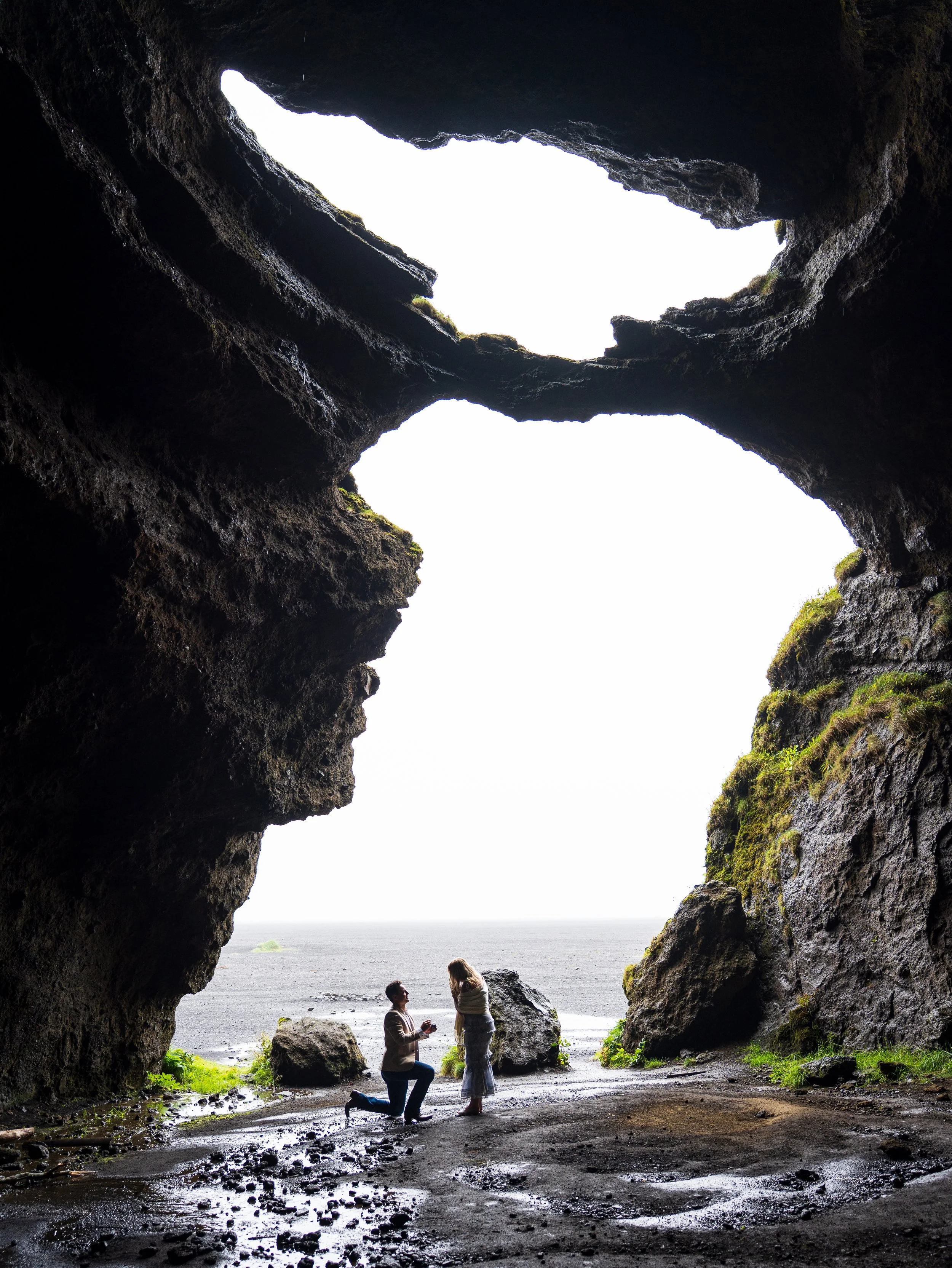 A couple proposing inside a rocky cave by the sea, with large rocks outside and ocean in the background.