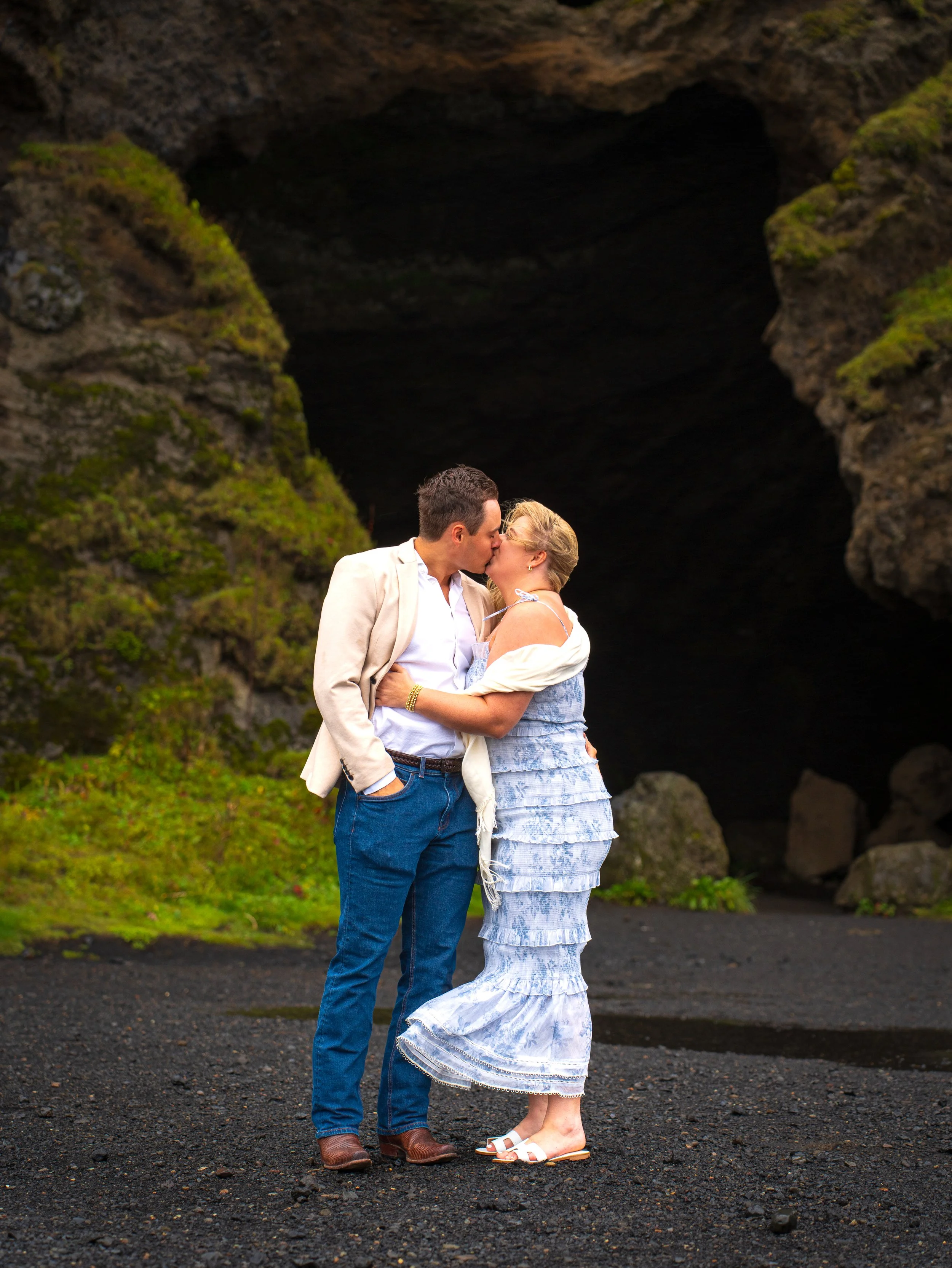 A couple kissing in front of a cave entrance, with rocky and mossy surroundings.