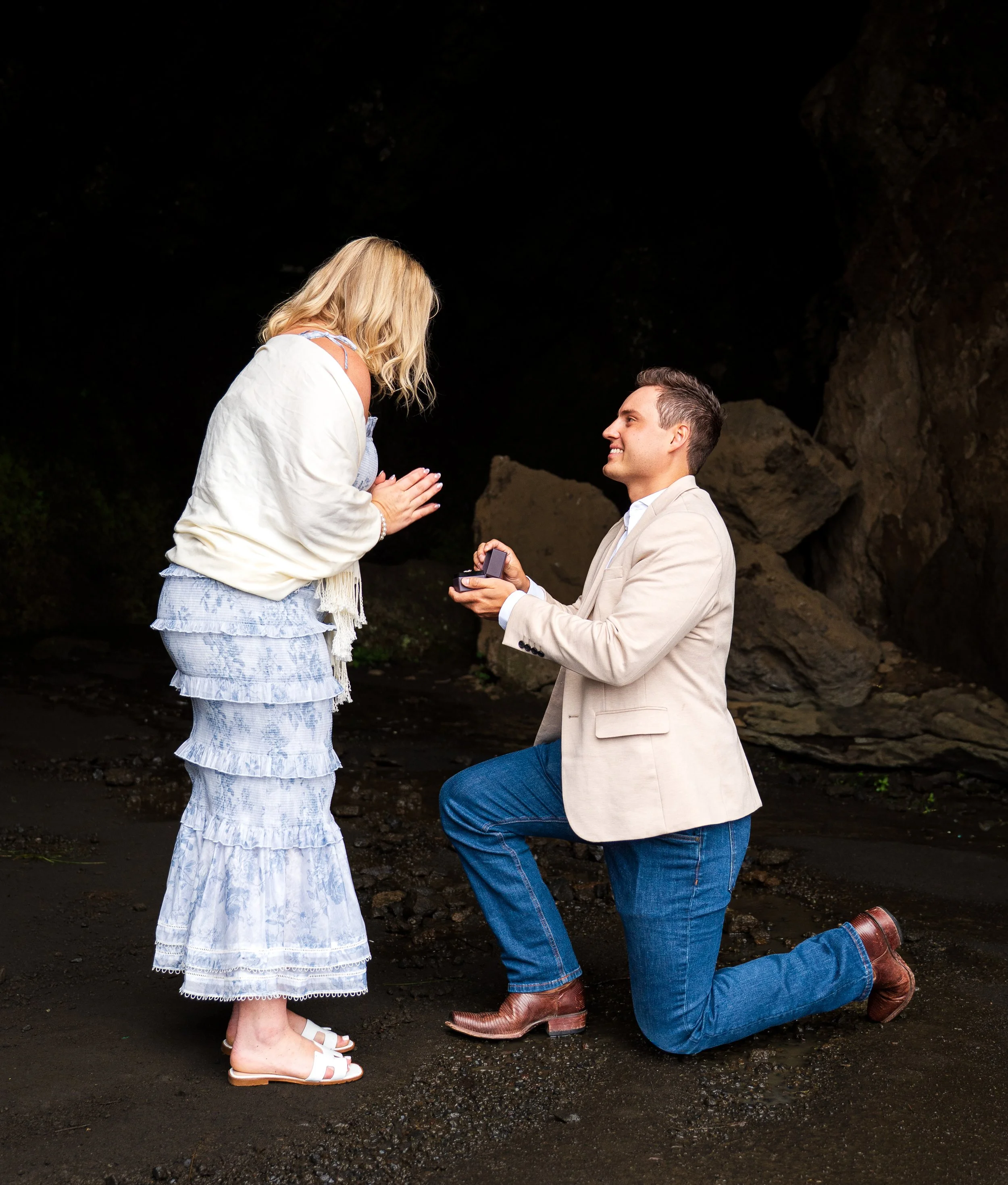 A man kneeling on one knee proposes to a woman inside a dark cave. The man is holding out a small box, likely with a ring, while the woman is surprised and clapping her hands. The woman is wearing a white and blue tiered dress, and the man is dressed