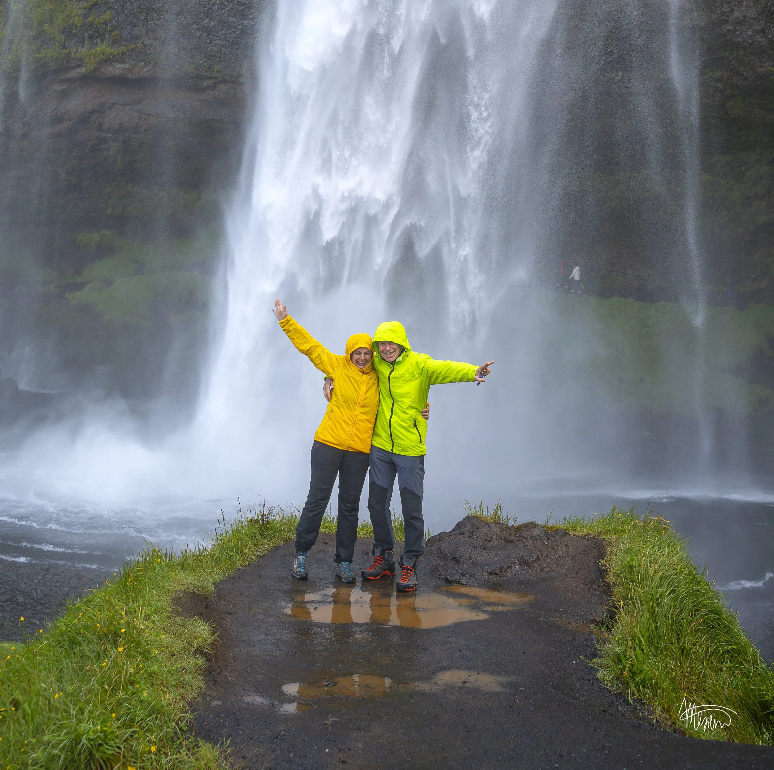 Two smiling people in yellow and green rain jackets standing on wet ground with a waterfall in the background, posing happily with arms around each other.