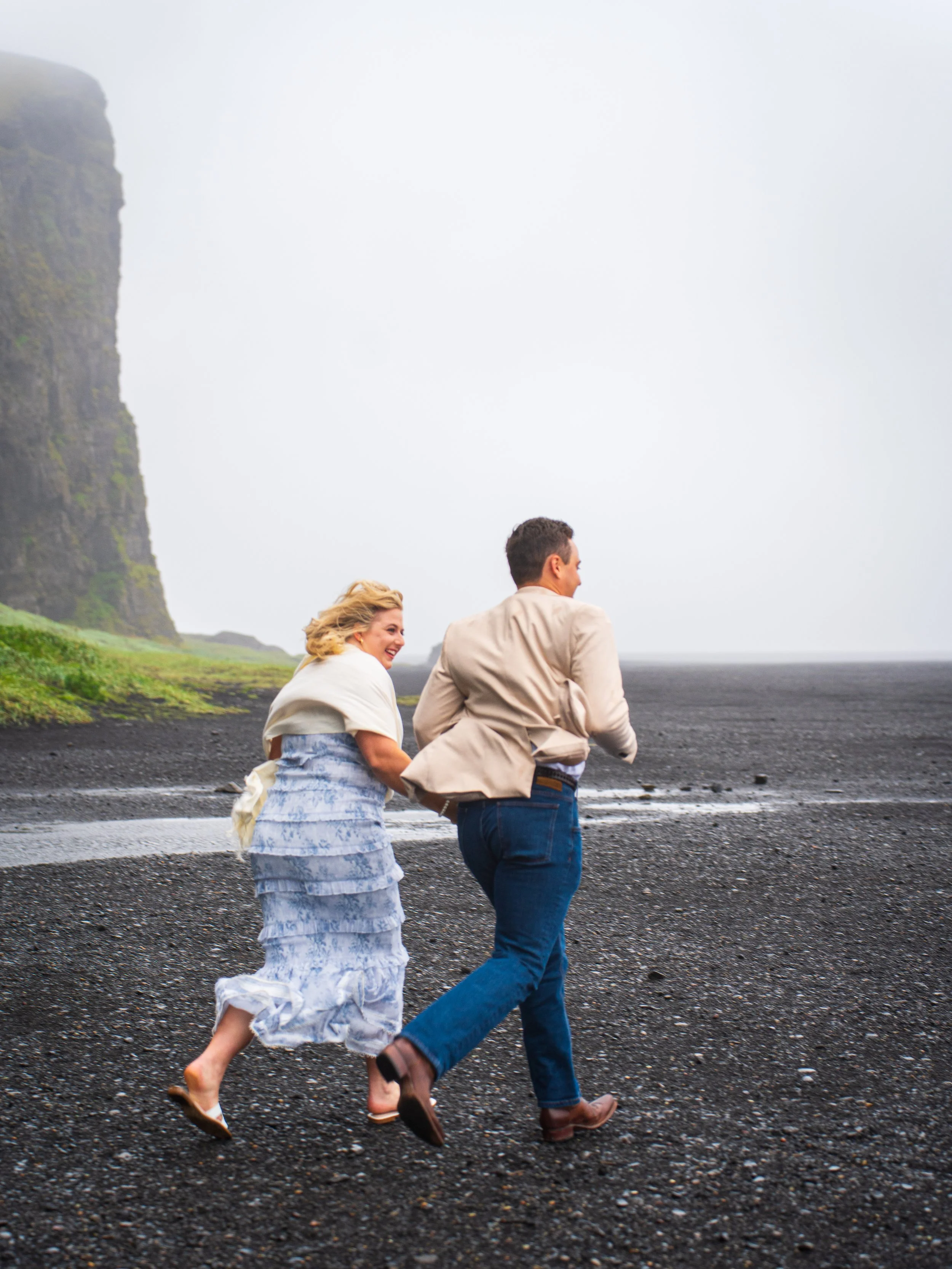 A couple is running on a pebbled beach, with the woman laughing and holding the man's arm, against a backdrop of towering cliffs and a cloudy sky.
