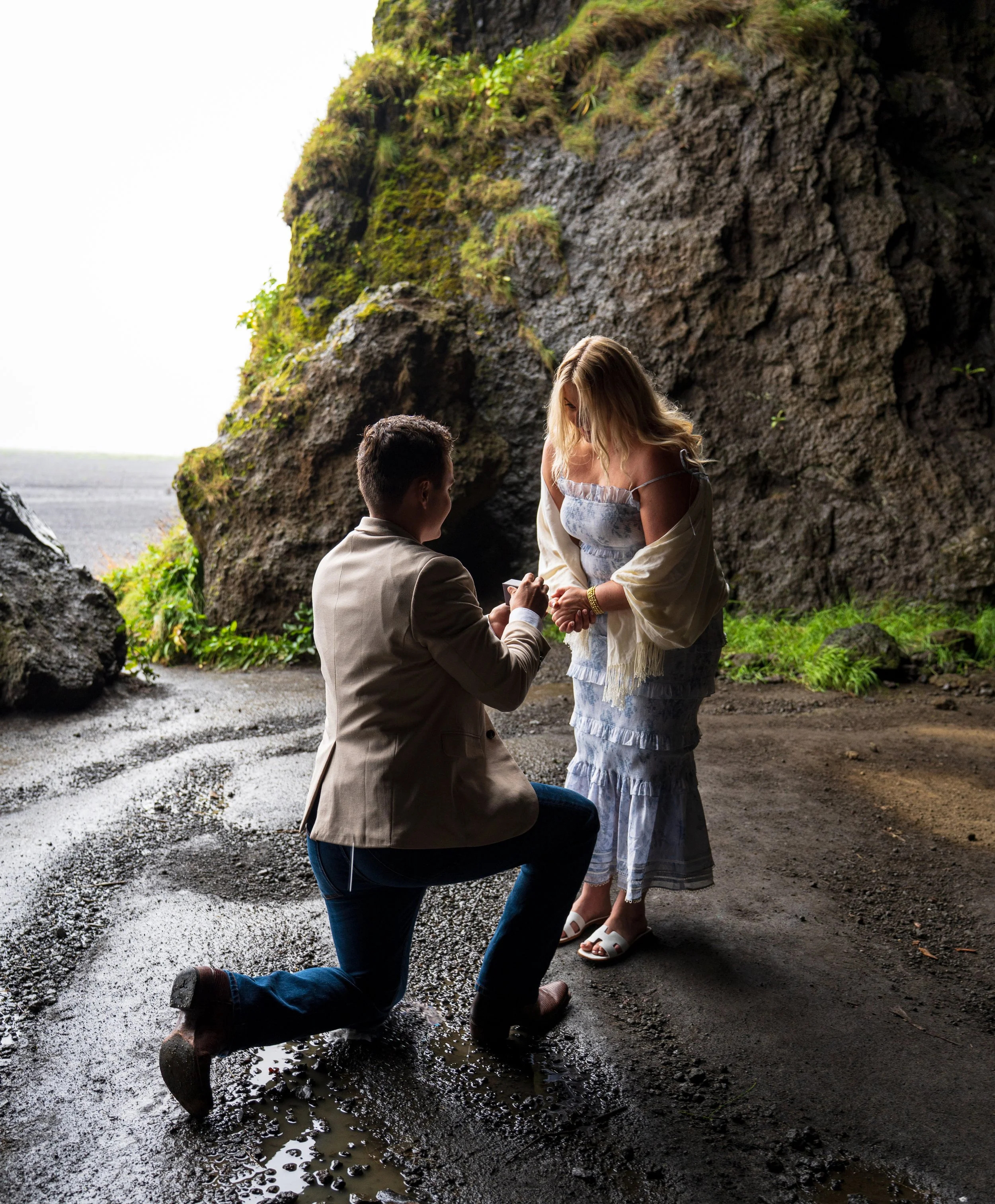 A kneeling man proposing marriage to a woman inside a cave with rocky walls and lush greenery, with the man kneeling on one knee holding her hand during a secret engagement photoshoot. Image taken and edited professionally by a local Photographer.