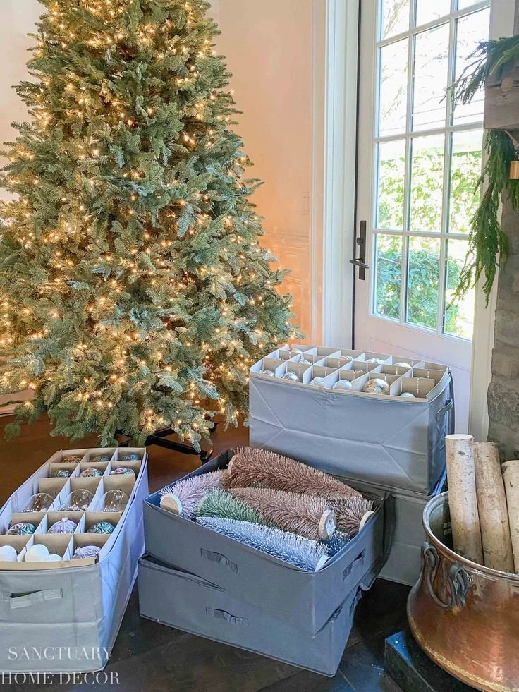 Decorative DIY Christmas ornaments in storage boxes near a decorated Christmas tree with lights, next to a window and a container with logs.