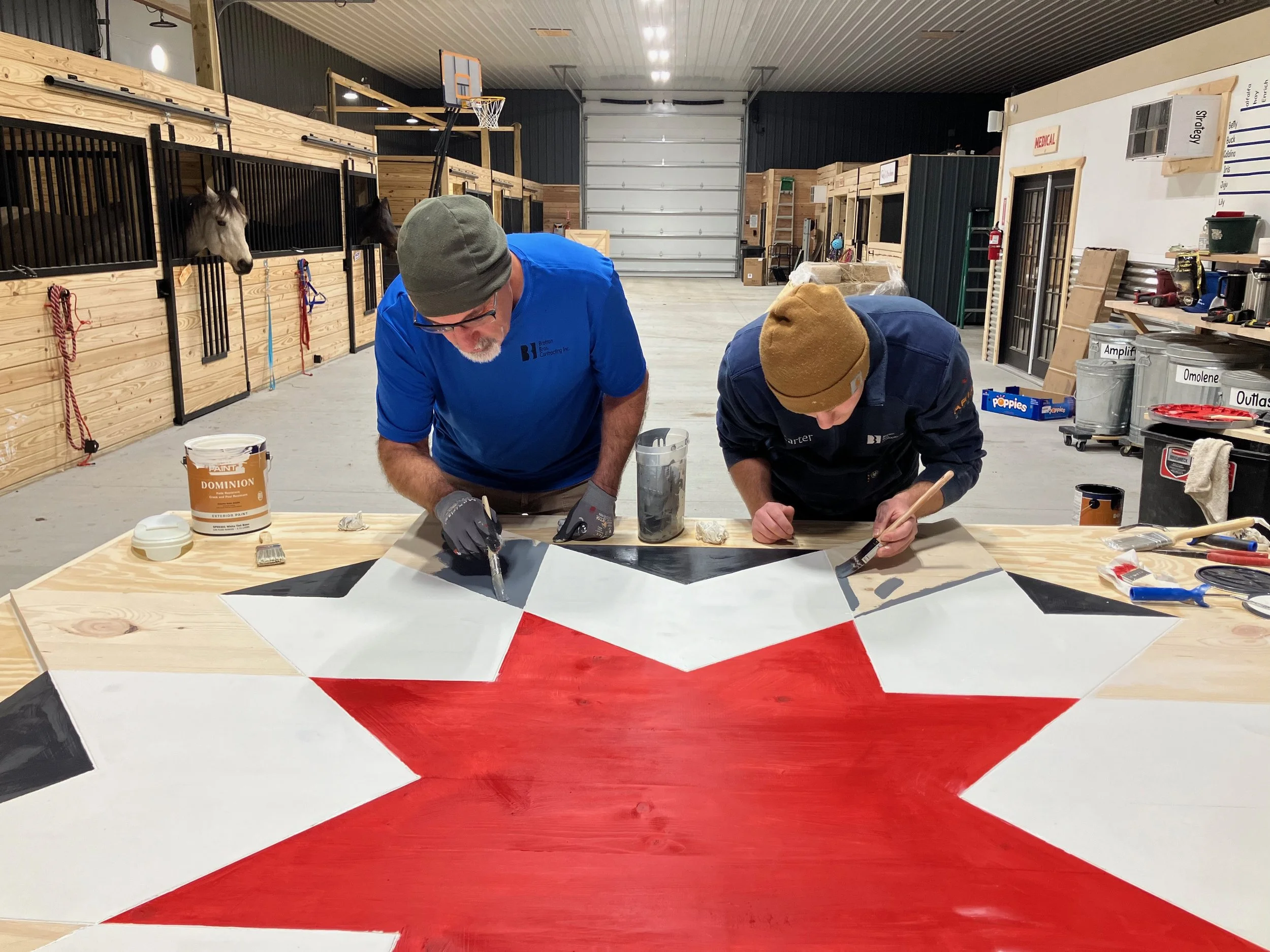 Two people painting a large wooden surface with geometric patterns in a barn, with horses visible in stalls. Paint supplies are on a table.