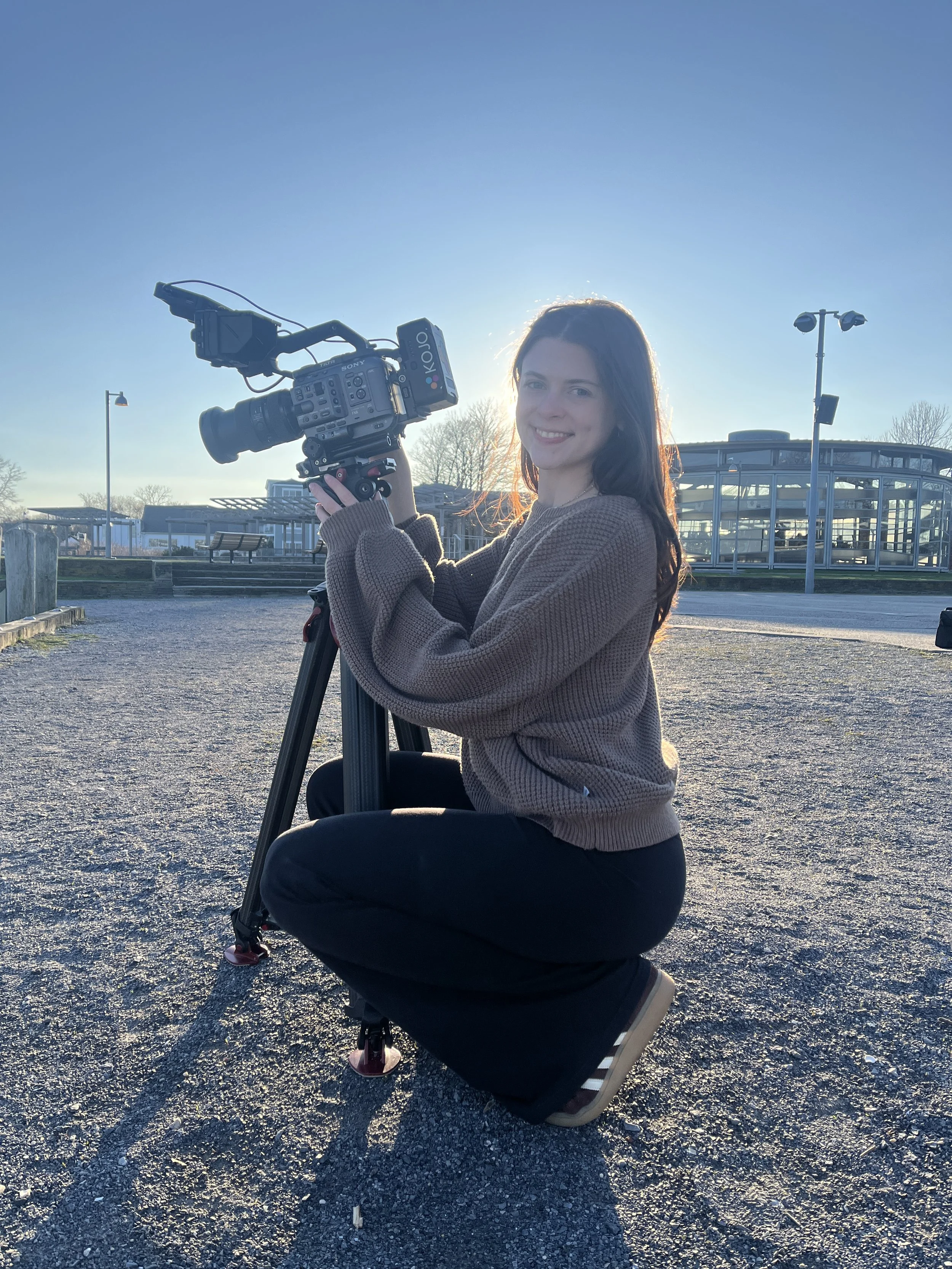 A woman is kneeling on gravel while holding a professional video camera mounted on a tripod. She is outdoors on a sunny day with the sun in the background, illuminating her face. The background includes modern glass buildings, trees, and a clear blue sky.